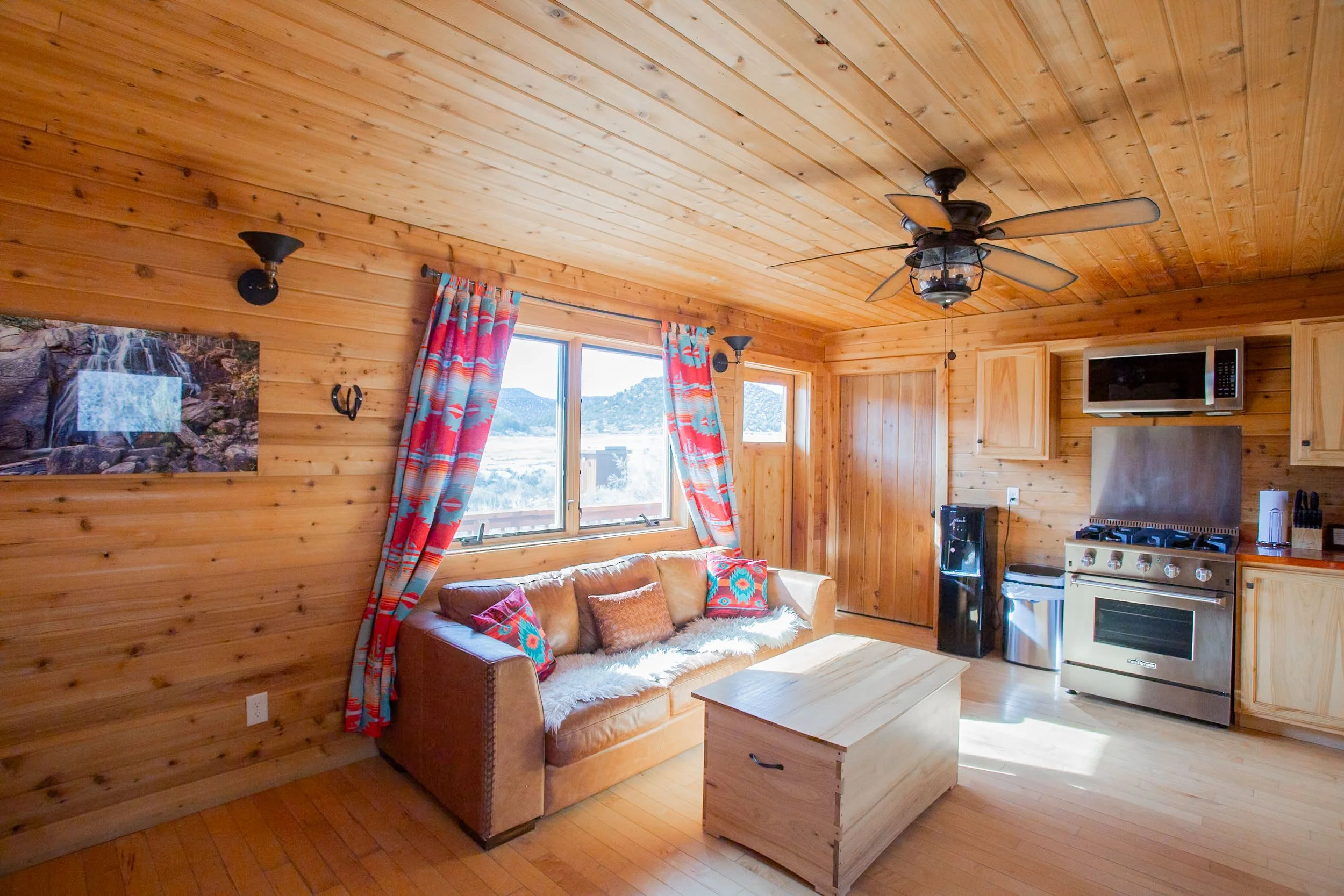 Interior of a cozy wooden cabin with a ceiling fan, a colorful curtain, a window showing a mountainous landscape, a leather couch with pillows, a wall-mounted TV, and a kitchen area with a stove and microwave.