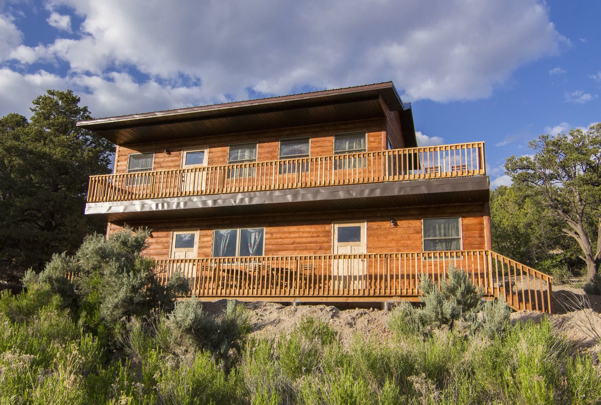 A wooden multi-story house with a balcony on the top floor, surrounded by trees and bushes under a partly cloudy sky.