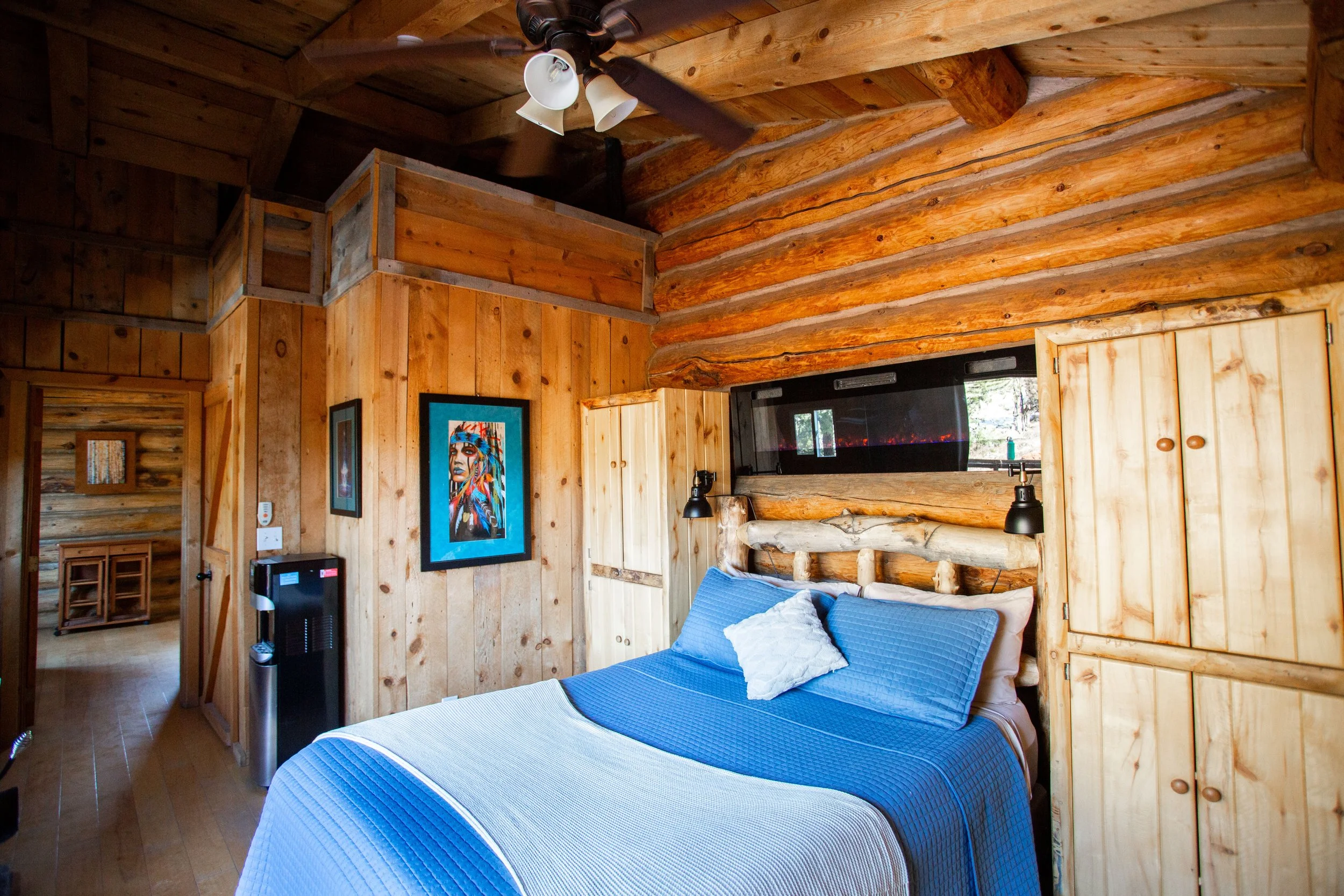 A cozy bedroom in a log cabin with wood walls and ceiling, featuring a bed with a blue quilt, a decorative pillow, and a wooden privacy screen or headboard. The room has black wall-mounted lamps, a colorful framed art piece, and a ceiling fan with lights.