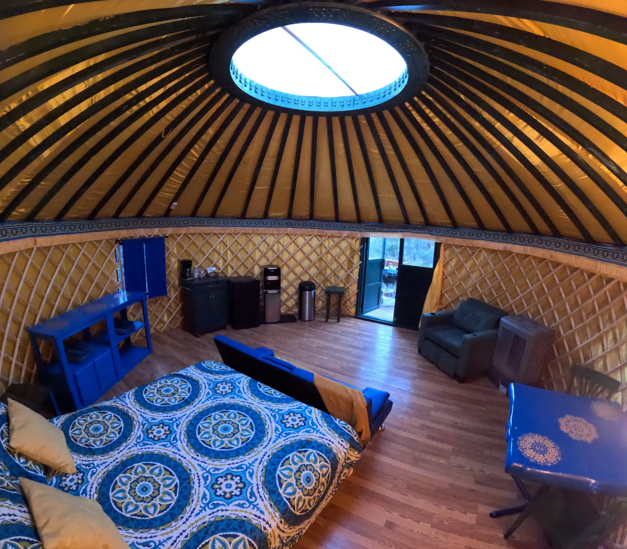Interior of a yurt with wooden lattice walls, a circular skylight, and furniture including a patterned bed, a blue bench, and a sofa.