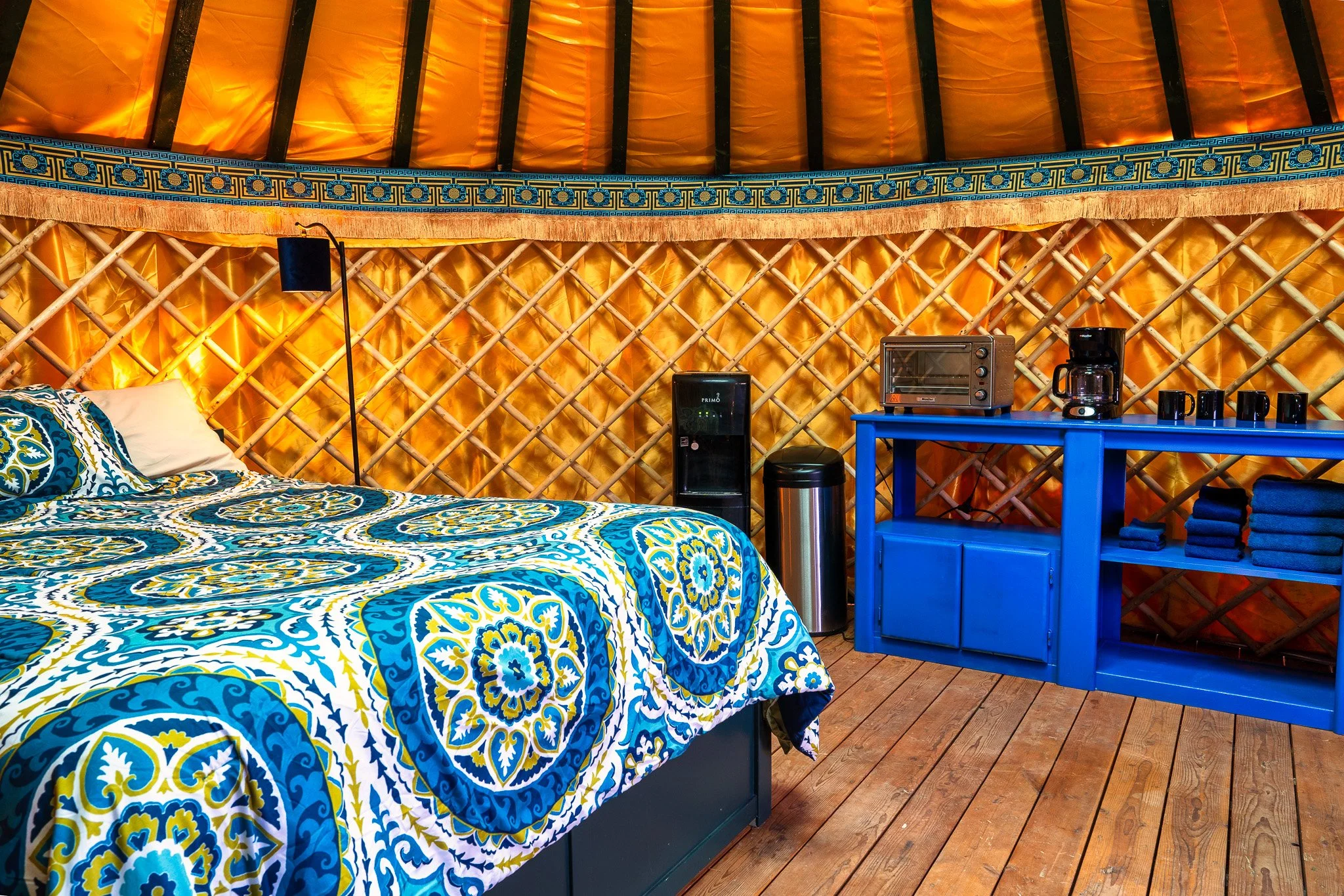 Interior of a bedroom with a bed covered in a blue and yellow patterned bedspread. The head of the bed is against a wall made of lattice wood panels with yellow fabric behind them. There is a blue nightstand with rolled blue towels, a coffee maker, a toaster oven, and a water dispenser. The floor is wooden.