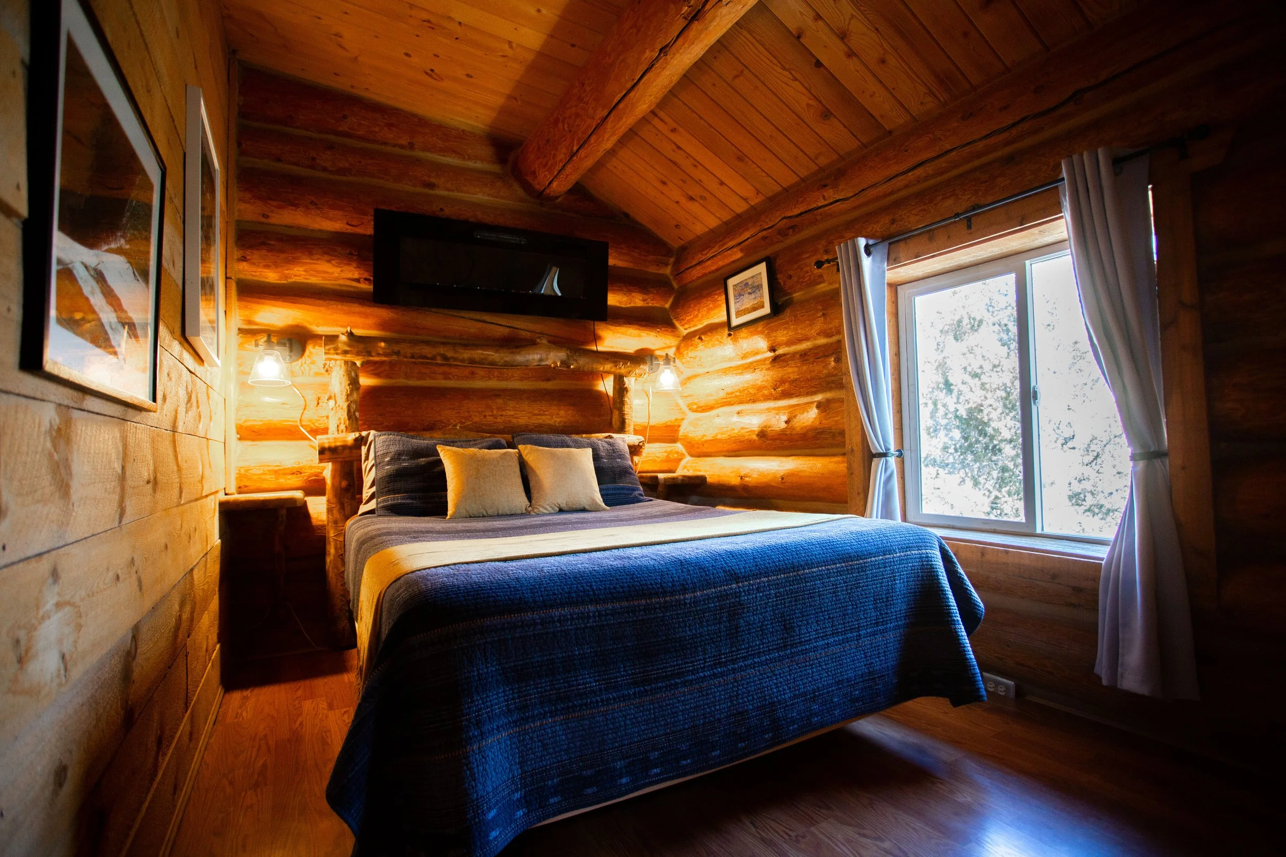 A cozy bedroom with wooden log cabin walls, a large window with curtains, a bed with pillows, and wall-mounted lamps.