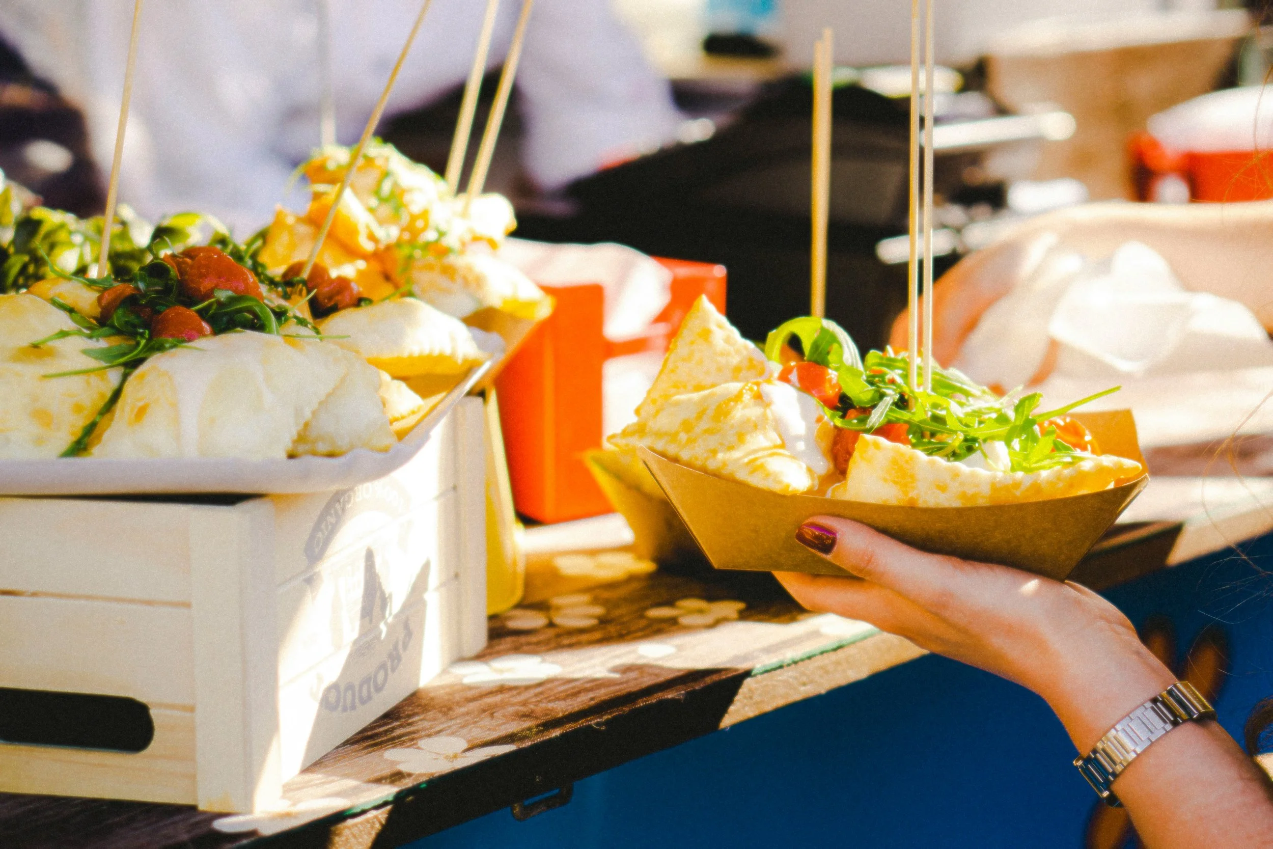 Person holding a paper cone filled with what appears to be food, with other food items and a box on a table, indicating a food or snack stand.