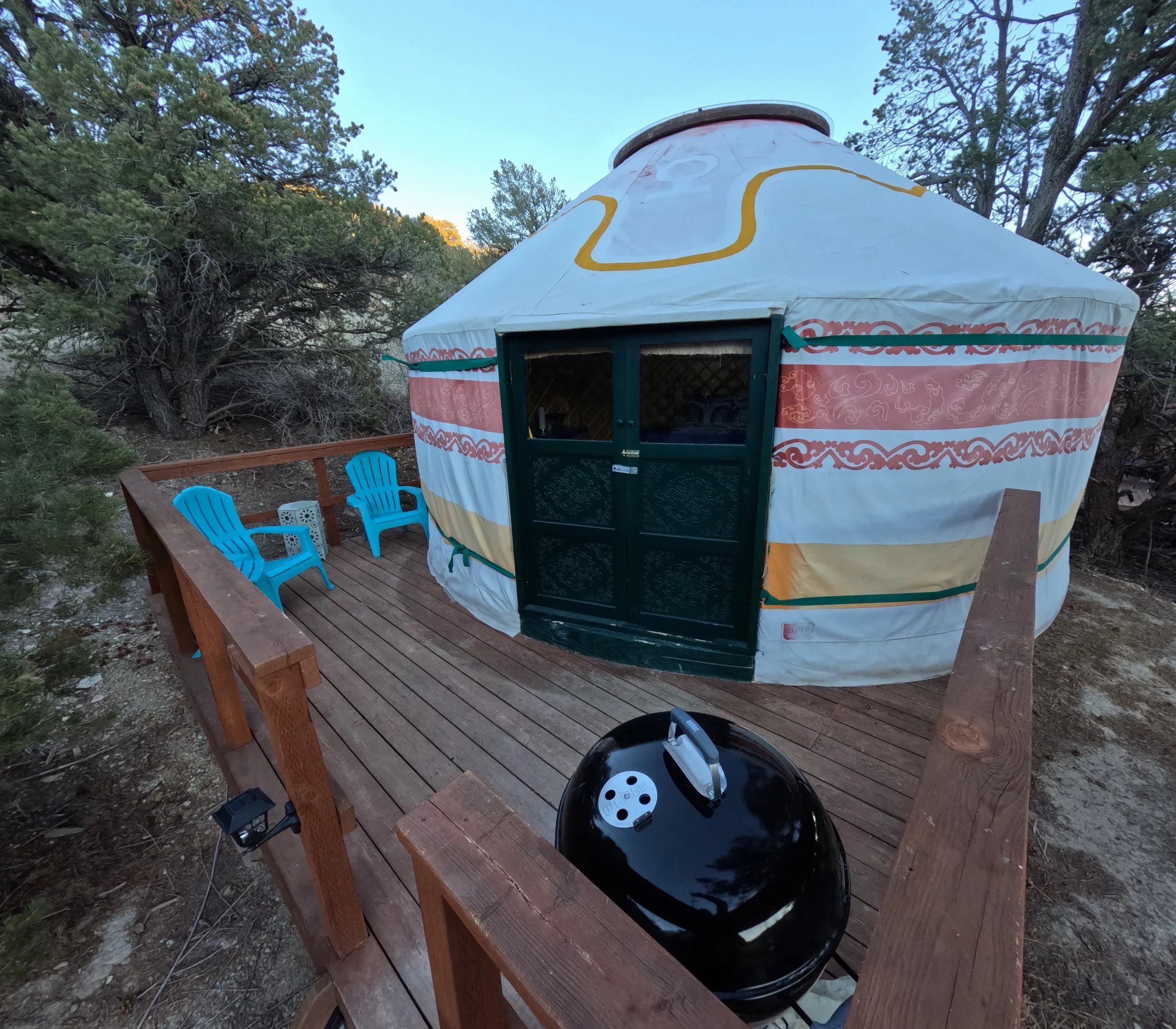 A campsite scene featuring a wooden deck with two blue chairs, a yurt-style tent, and two motorcycle helmets, with trees and a clear sky in the background.