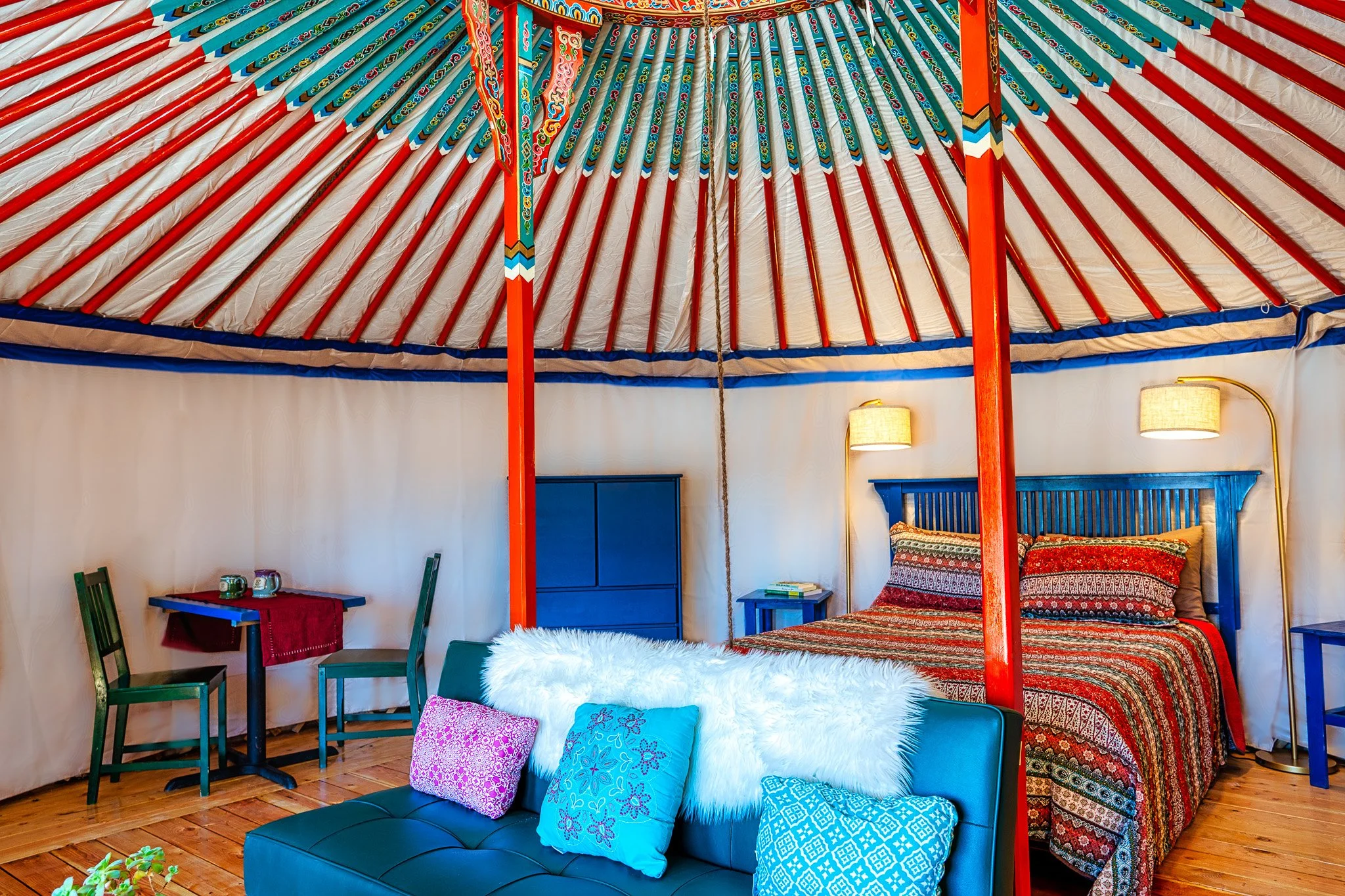 Interior of a room featuring a colorful, decorative canopy bed with red, orange, and blue painted poles, and patterned bedding. There are walls with mounted lamps, a small table with green chairs, a fluffy white chair with pillows, and a wooden floor.