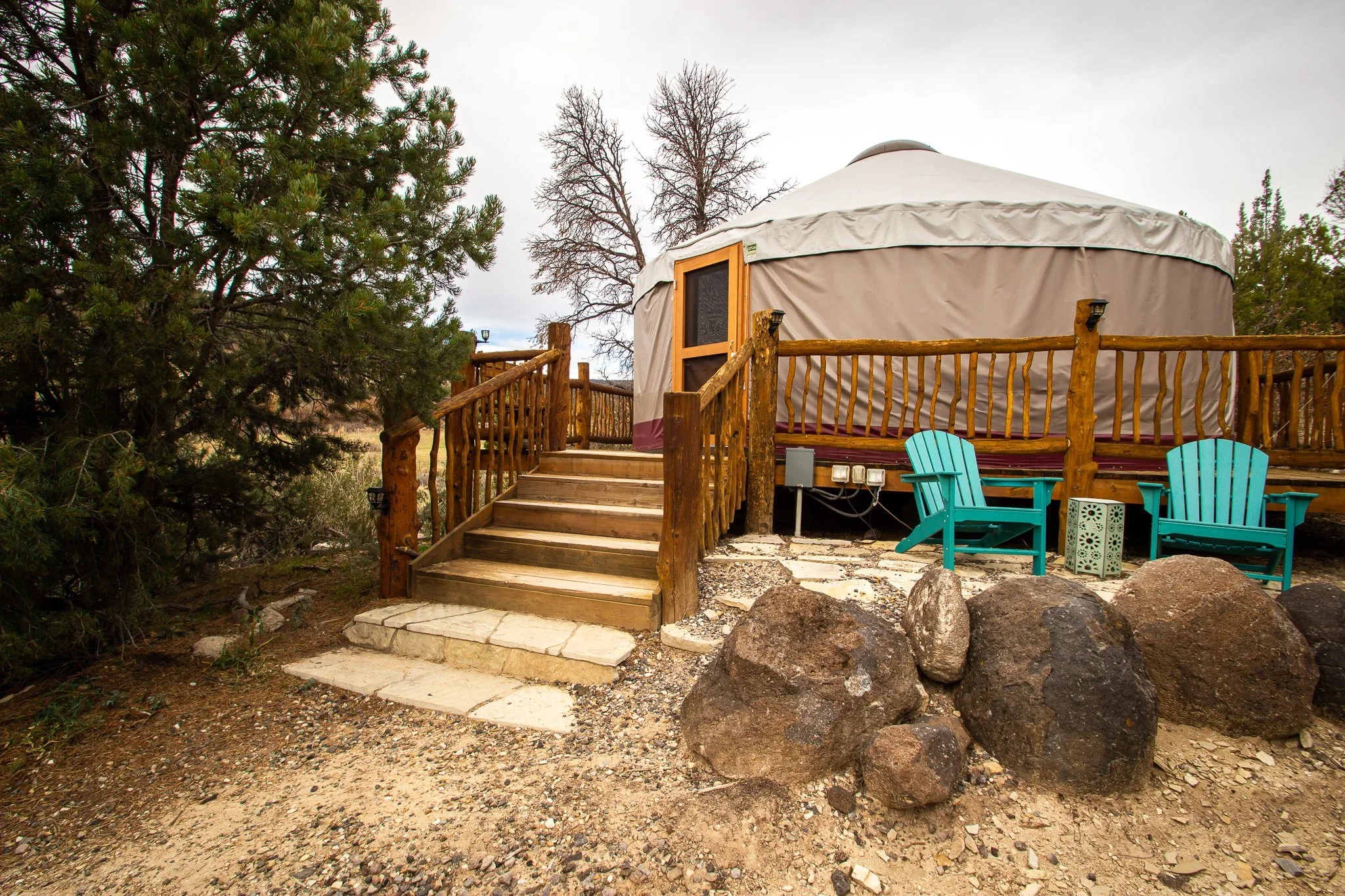 A yurt with a wooden deck and stairs, surrounded by some trees and rocks, with two colorful Adirondack chairs outside.