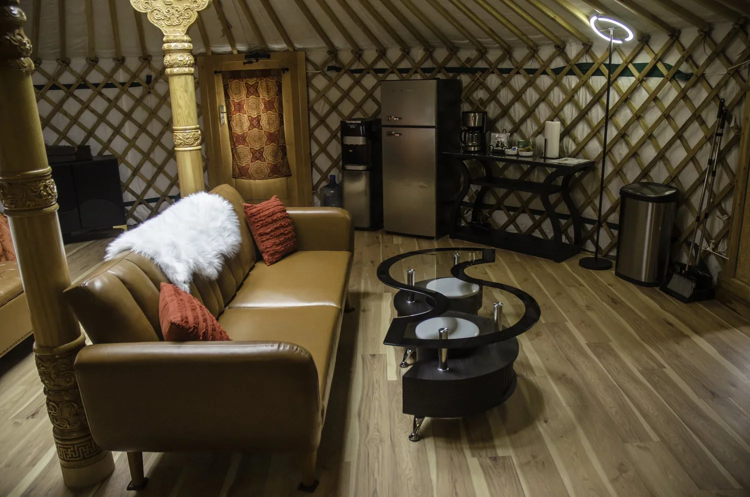 Interior of a cozy yurt-style lounge with wooden lattice walls, a leather couch with red pillows and a white furry throw, a unique black coffee table, a small bar with a refrigerator and coffee maker, and a neon heart-shaped light.