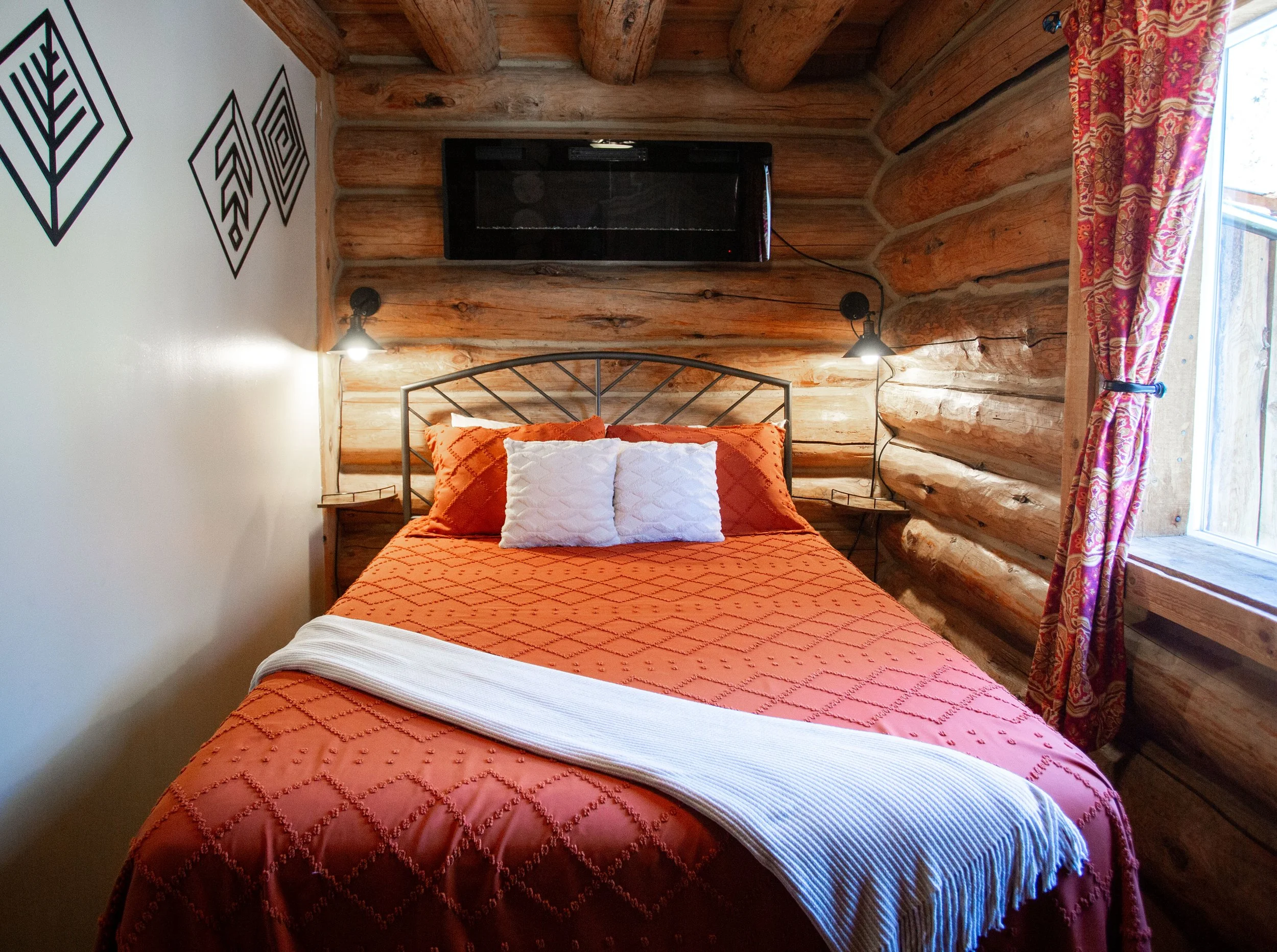 A cozy bedroom with log cabin walls, a bed with an orange quilt, white and orange pillows, wall-mounted reading lamps, a flat-screen TV, and a window with patterned pink curtains.
