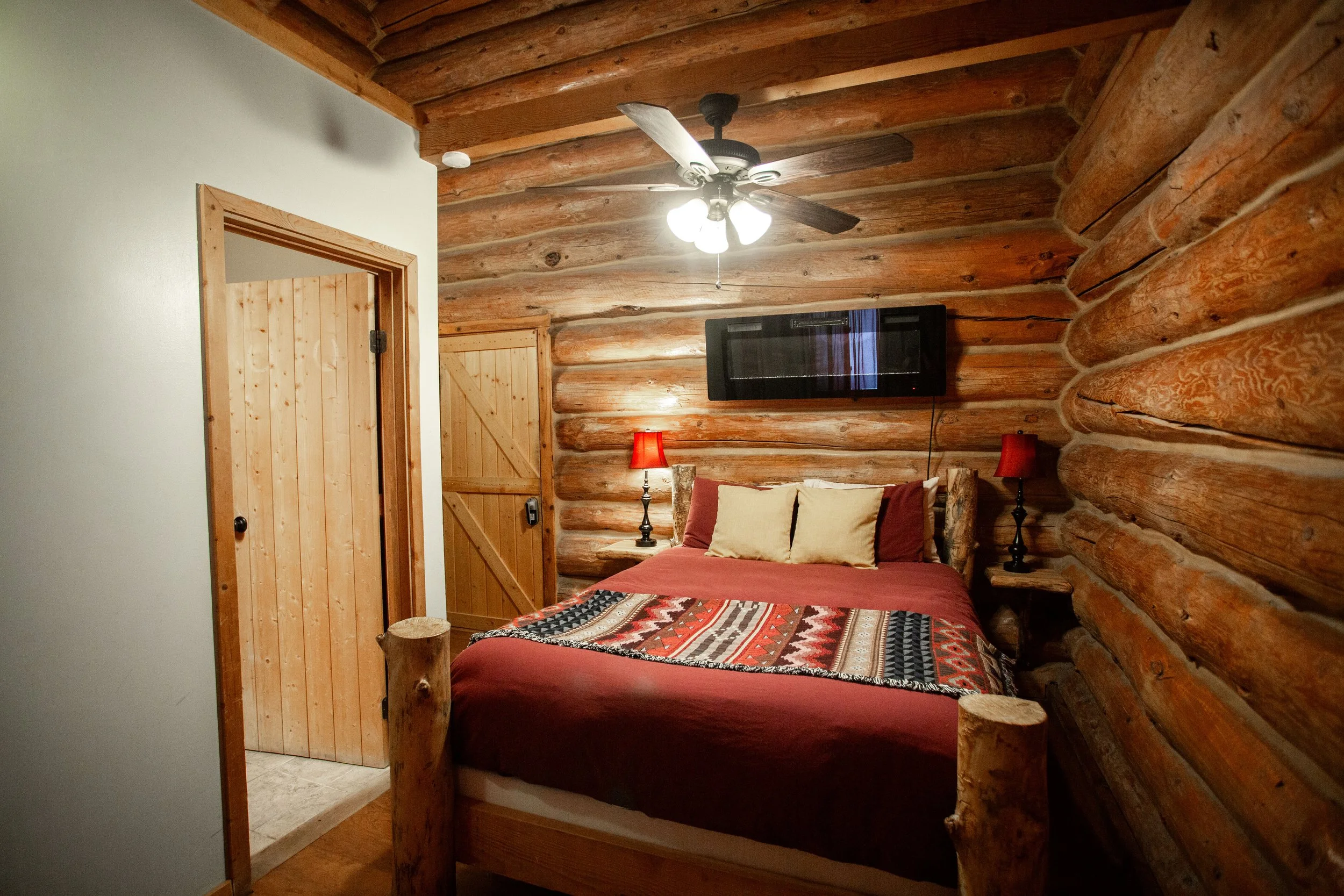 A cozy rustic bedroom with log walls, a wooden ceiling with a fan, a wall-mounted TV, a bed with a decorative quilt, two red lamps on matching side tables, and a wooden door.