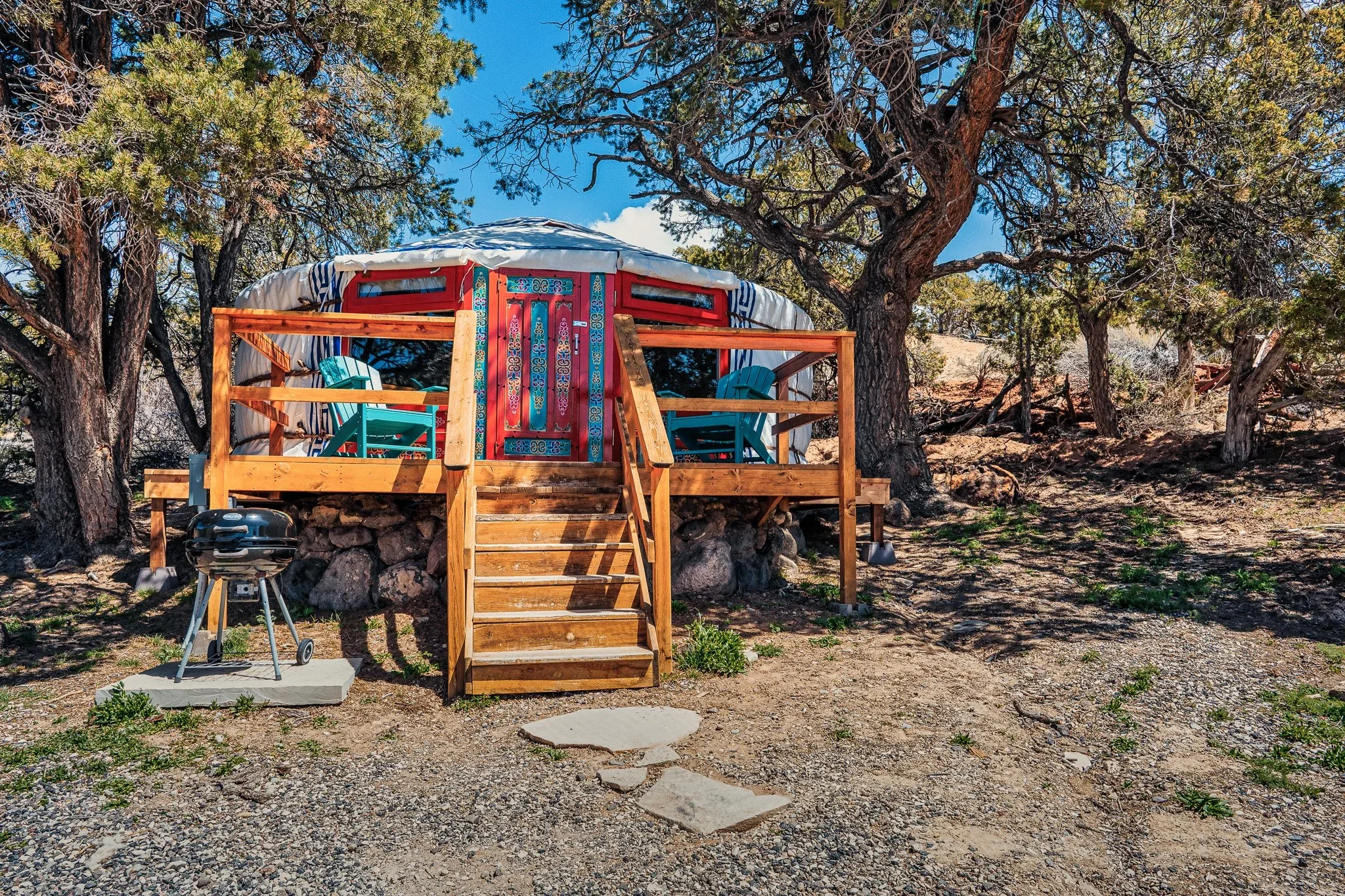 A colorful yurt with a red door and decorated patterns, situated on a wooden deck with blue chairs, surrounded by trees in a natural outdoor setting.