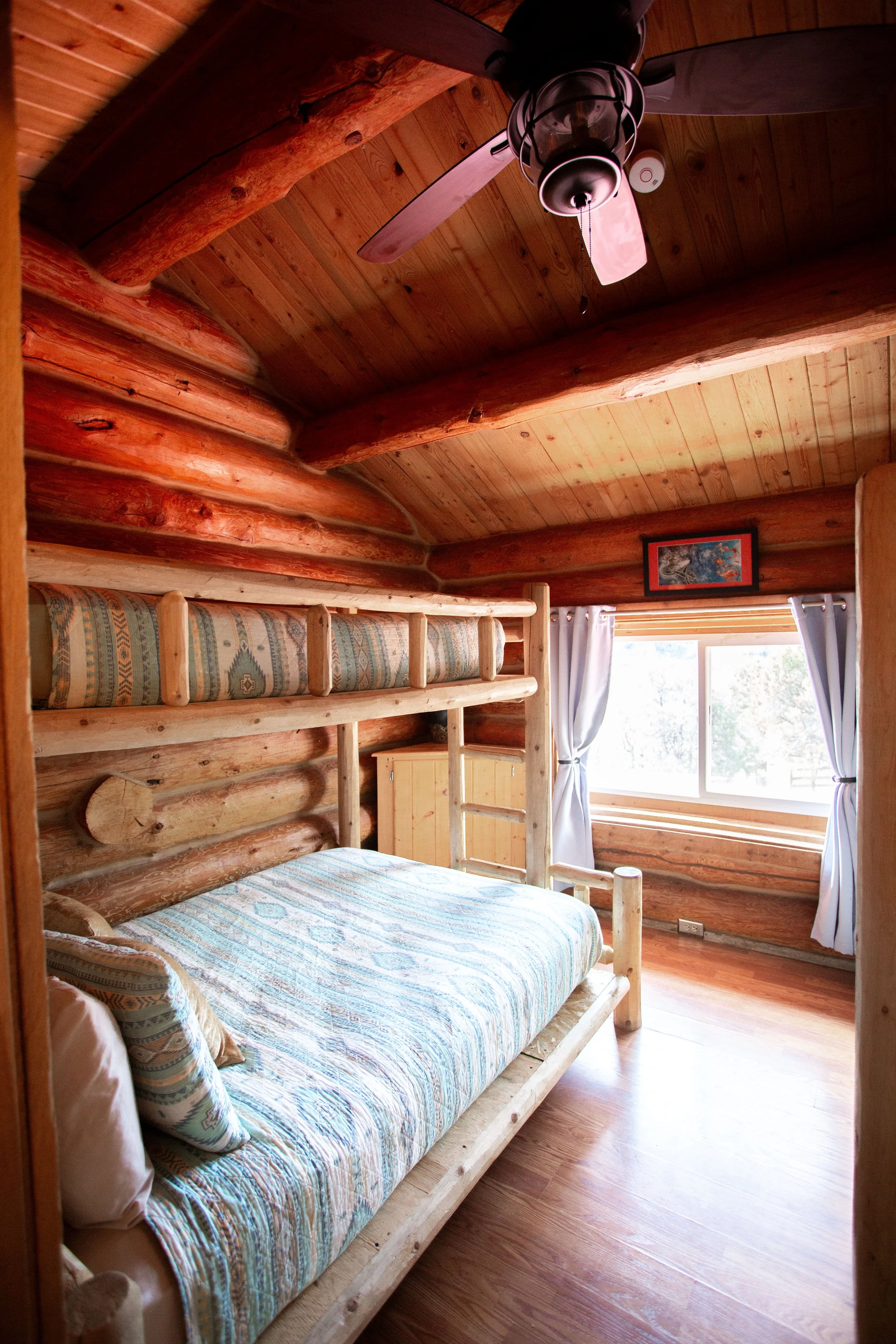 A rustic bedroom with a wooden log bed frame and a patterned mattress, large window with white curtains, and wooden ceiling with a ceiling fan.