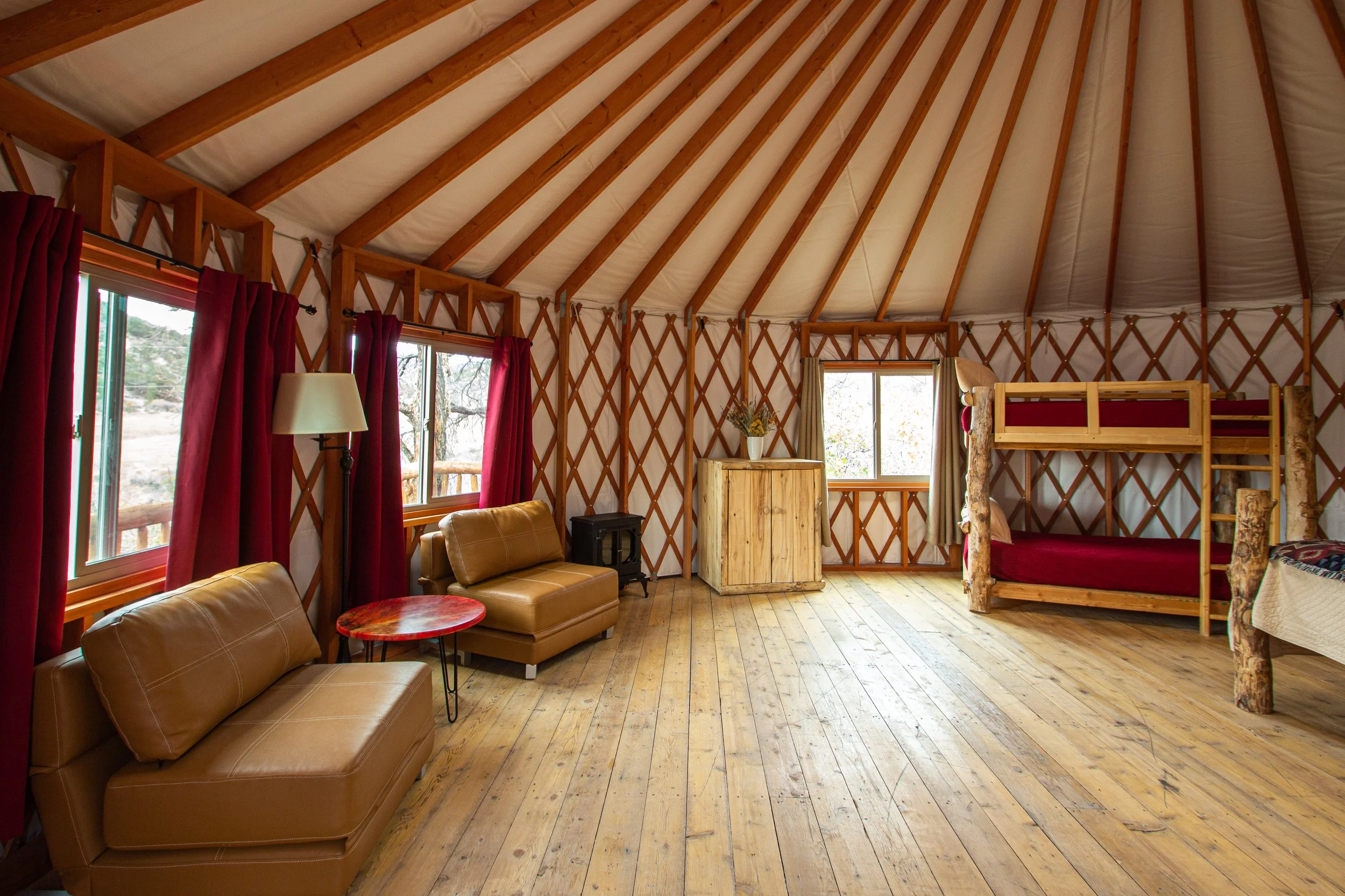 Interior of a cozy wooden cabin with natural wood floors, red curtains, leather sofas, a wooden dresser, and a loft bed. Large windows reveal trees outside.