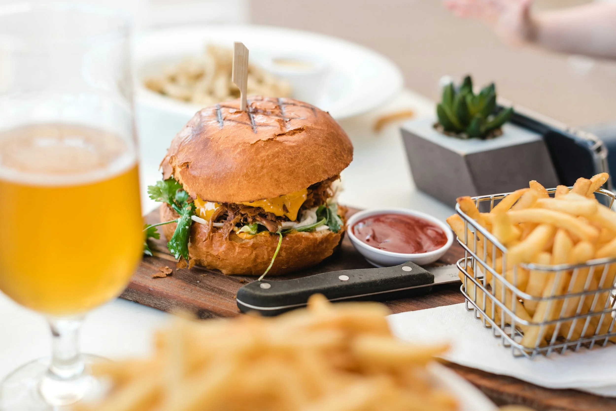 A hamburger with lettuce, cheese, and shredded meat on a bun, served with ketchup, French fries in a wire basket, a glass of beer, and a side dish of pasta in the background, on a wooden serving board with a knife.