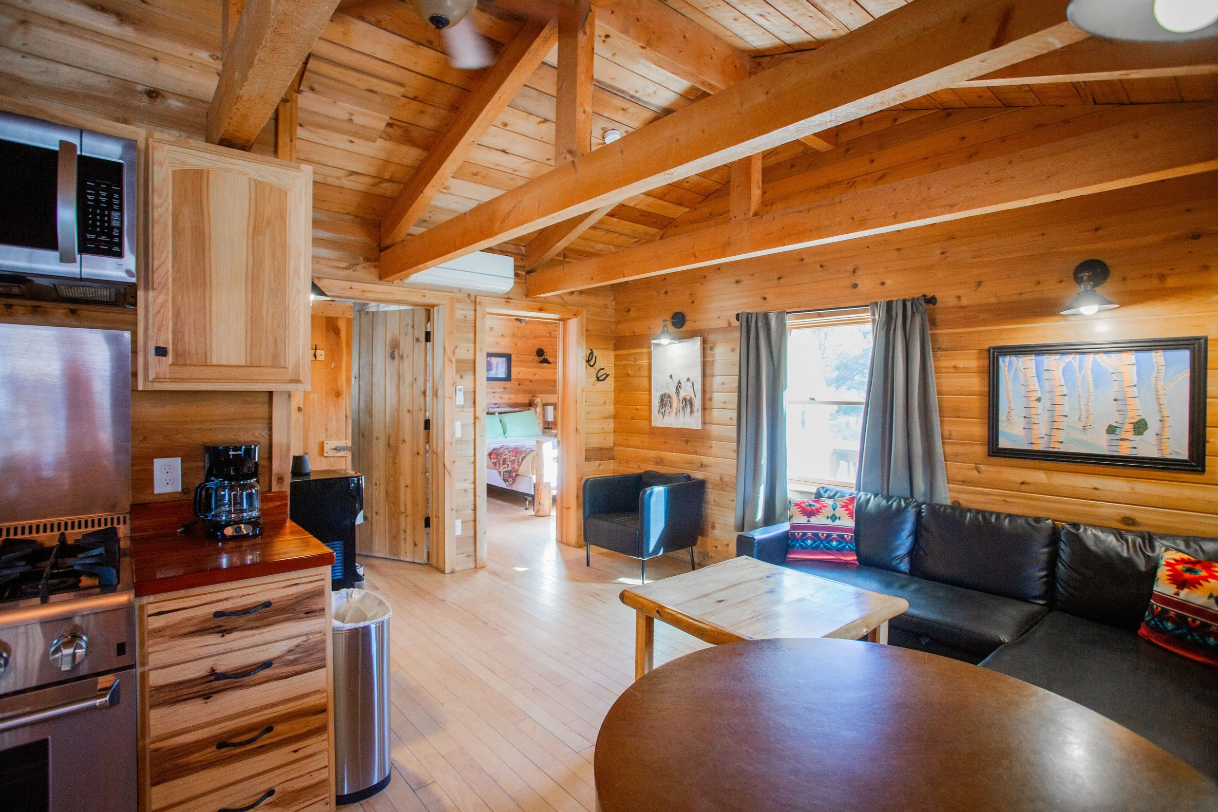 Interior of a cozy wooden cabin living room with wood-paneled walls and ceiling, a black leather sofa with colorful throw pillows, a window with blue curtains, and wall art. Part of a kitchen with stainless steel appliances and a coffee maker is visible.