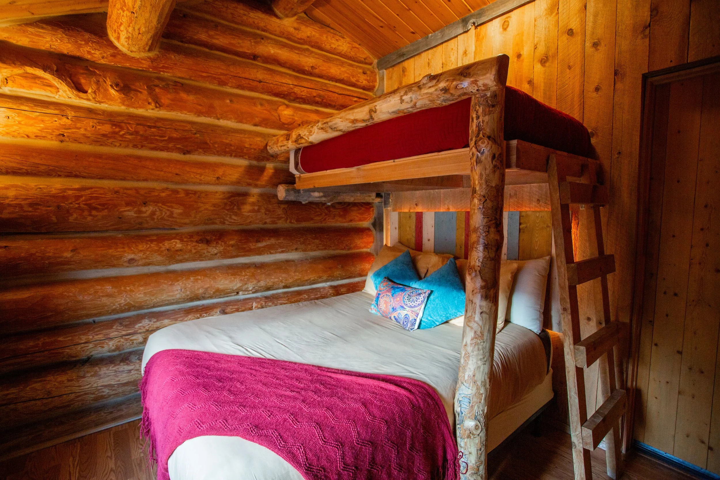 A cozy bedroom with log cabin-style decor, featuring a wooden bed frame and colorful pillows, with wooden paneling on the walls and ceiling.