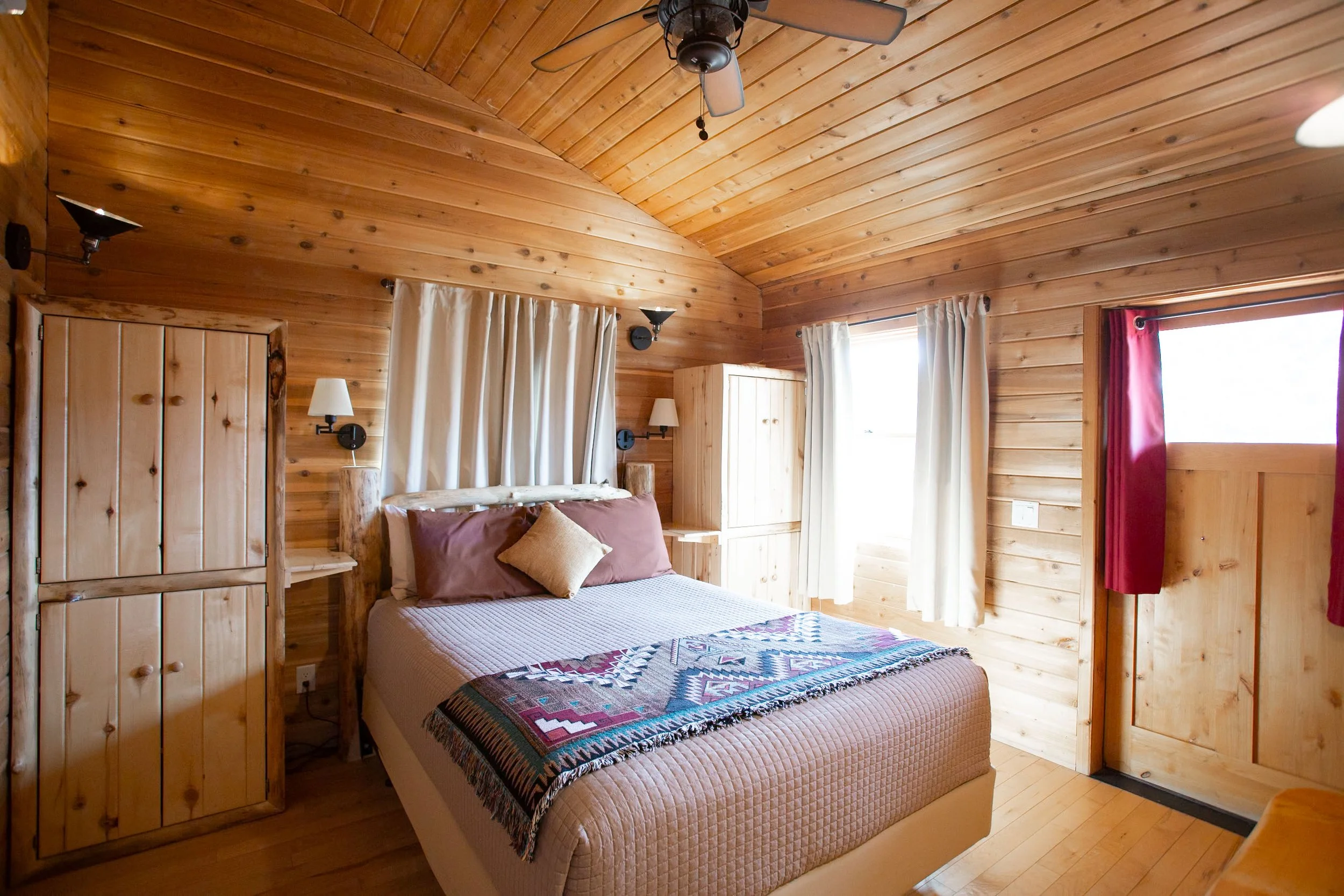 A cozy bedroom with wood-paneled walls and ceiling, a bed with a quilt and decorative pillows, wooden nightstands with lamps, and windows with white curtains and a red curtain.