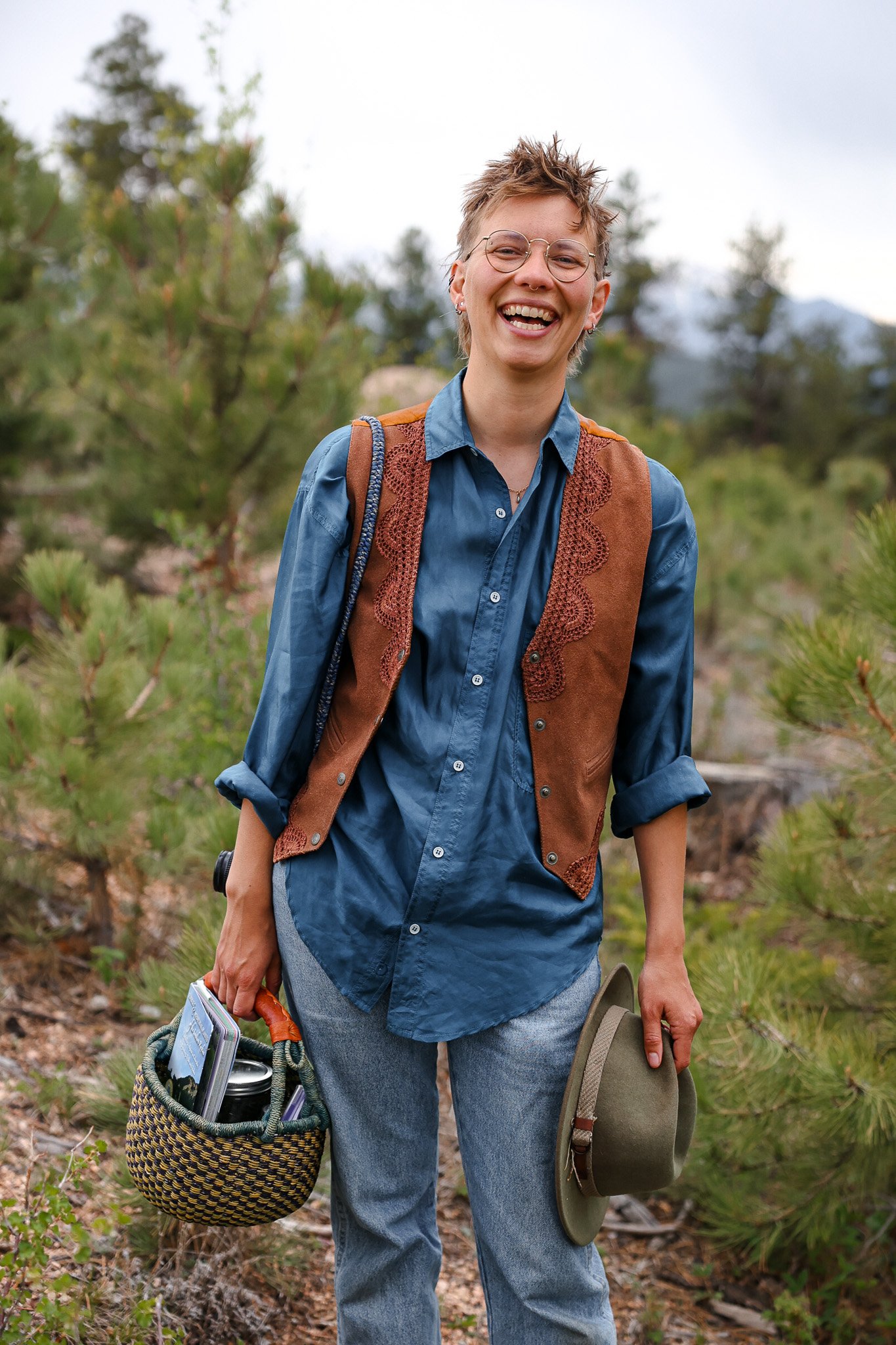 A woman smiling outdoors, wearing glasses, a blue shirt, a brown vest, and jeans, holding a hat in one hand and a woven basket with a book and a thermos in the other, in a forested area.
