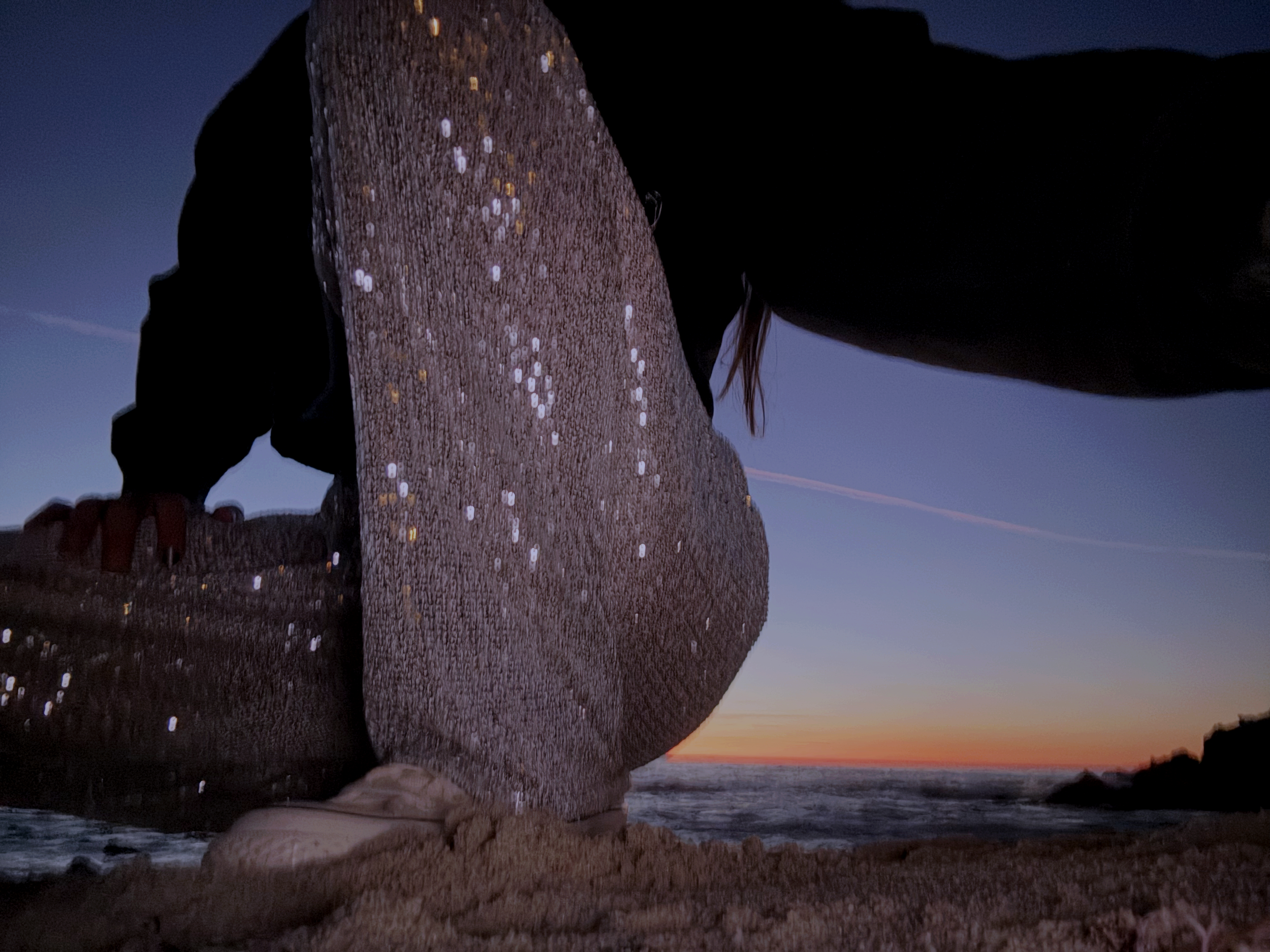 Person crouching on a rocky beach at sunset, with the ocean and colorful sky in the background. The person is wearing dark clothing and beige shoes, partially obscured by a large rock.