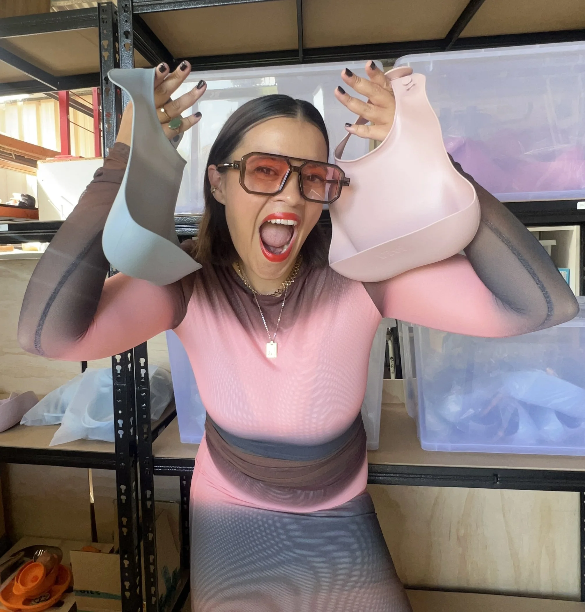 Woman with glasses and red lipstick holding two silicone bras, one gray and one pink, with an excited expression.
