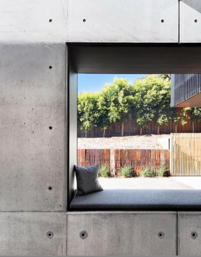 A built-in window seat with a gray cushion and pillow, looking out onto a backyard with a wooden fence, plants, and trees.