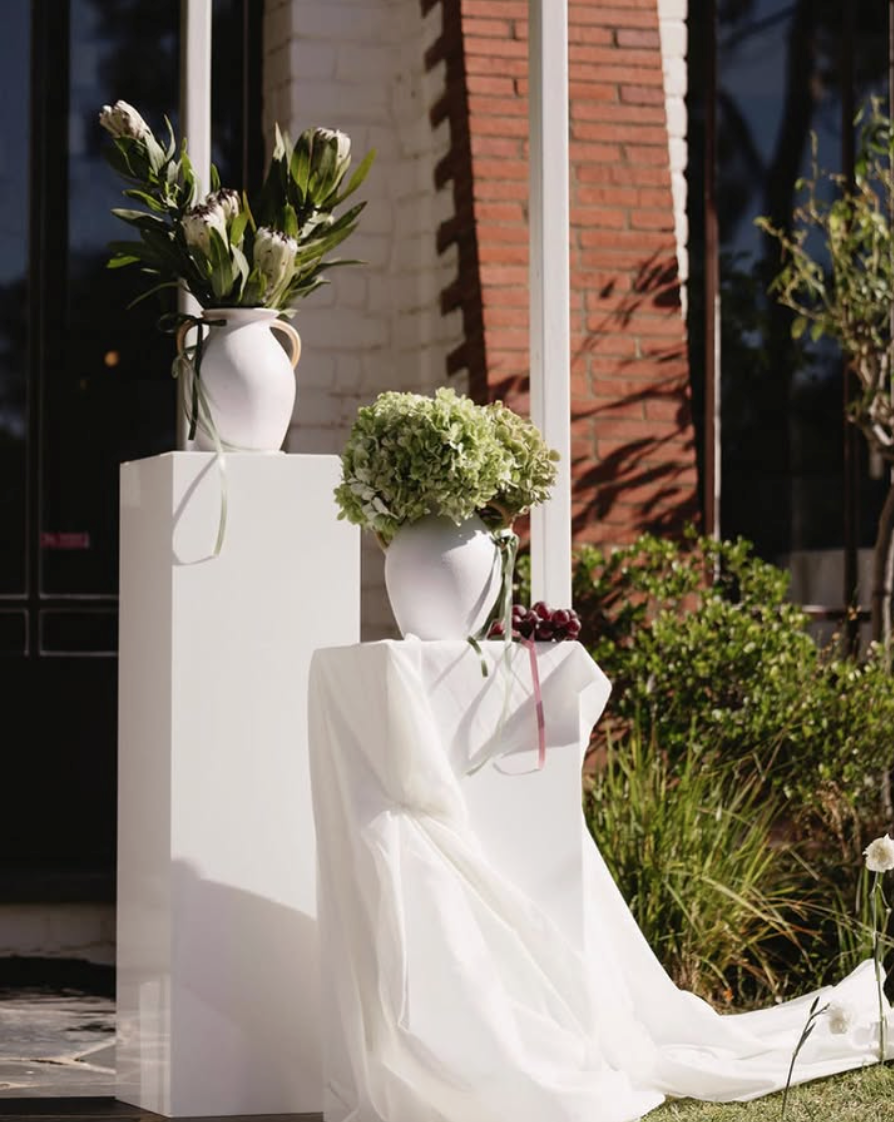 White vases with flowers on white pedestals outside near a brick and glass building, with a white draped cloth.