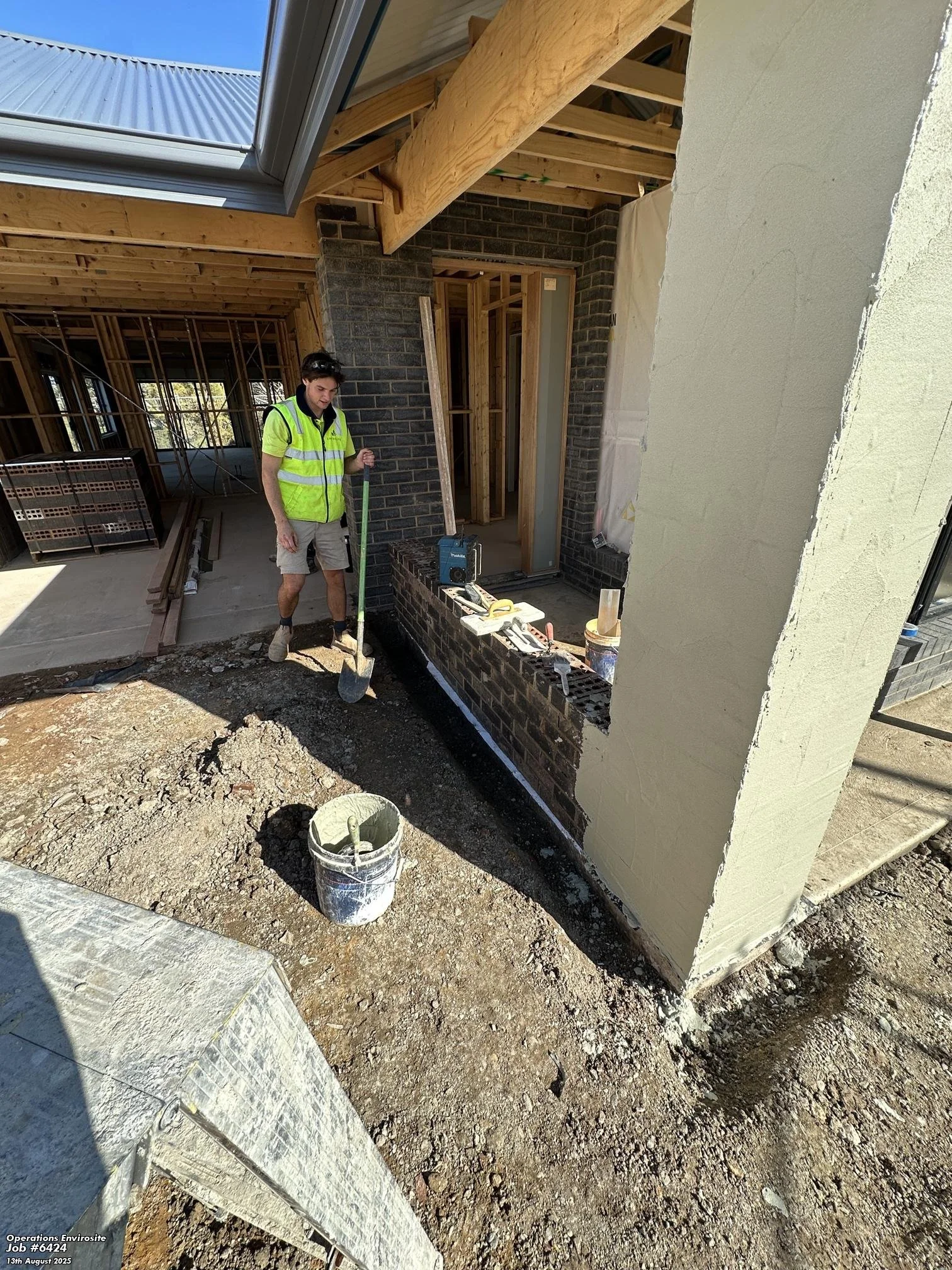 Construction worker in a neon green safety vest working on an outdoor brick wall, with exposed wooden roof structure and construction tools nearby.