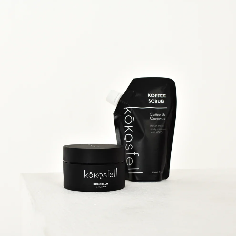 Black Koko Balm container labeled 'kökösfell KOKO BALM' and a black Kozisteil coffee scrub pouch labeled 'KOFFEE SCRUB Coffee & Coconut' on a white surface with a white background.