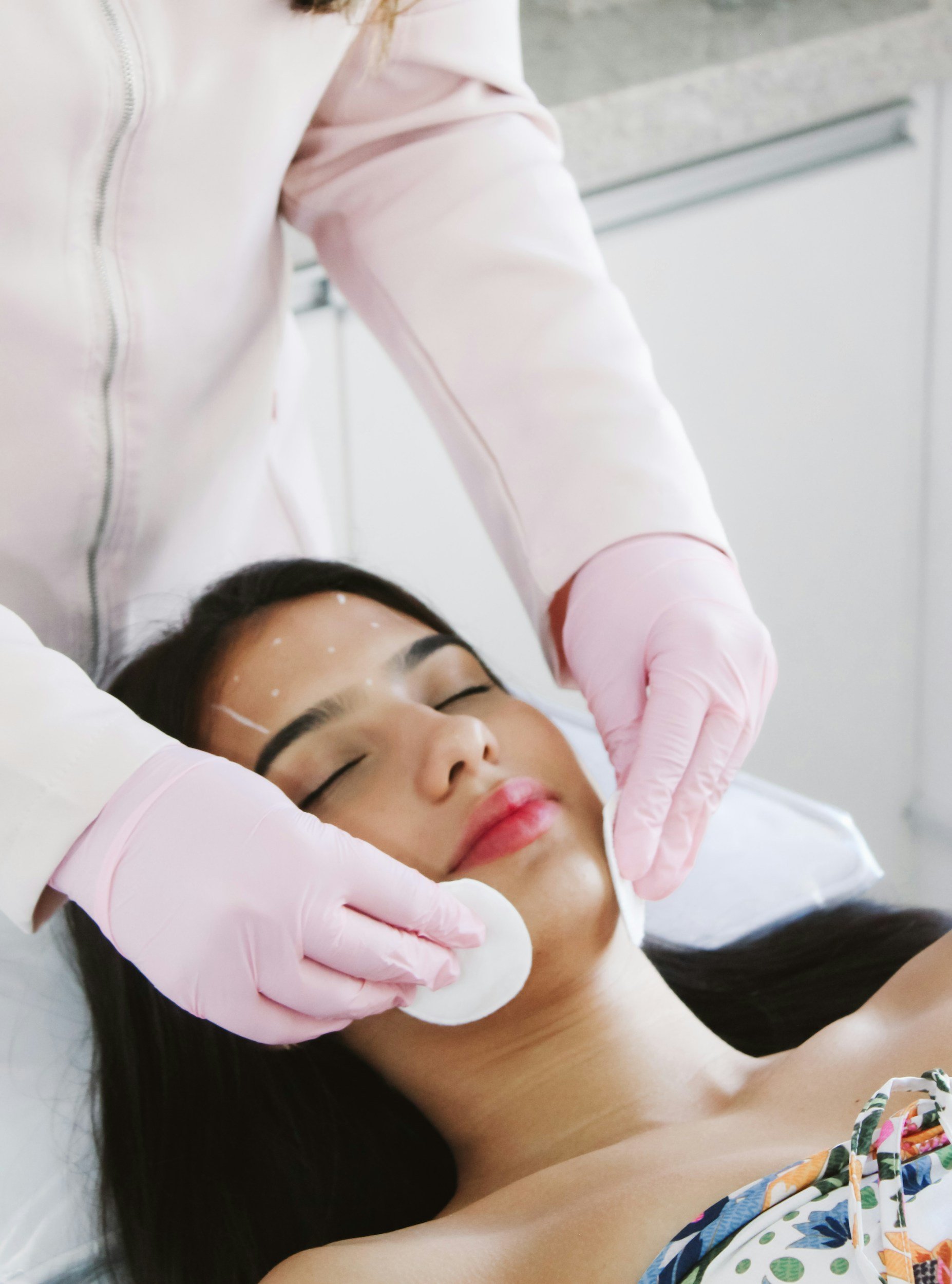 A woman lies down with eyes closed with white marks on her face while a person wearing pink gloves holds cotton pads, representing at home botox services