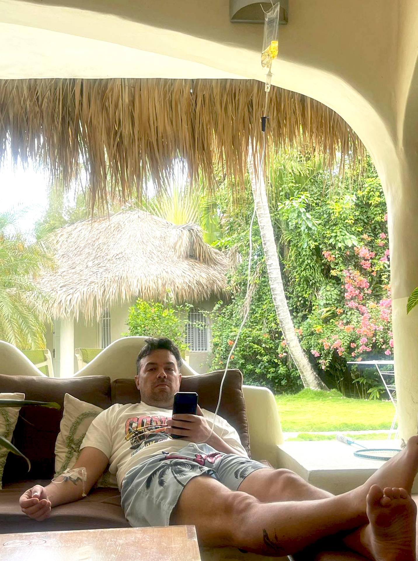 Man lying on a couch taking a selfie with a smartphone in a tropical outdoor setting with palm trees, flowering bushes, and a thatched roof structure visible outside. Receiving an Iv therapy