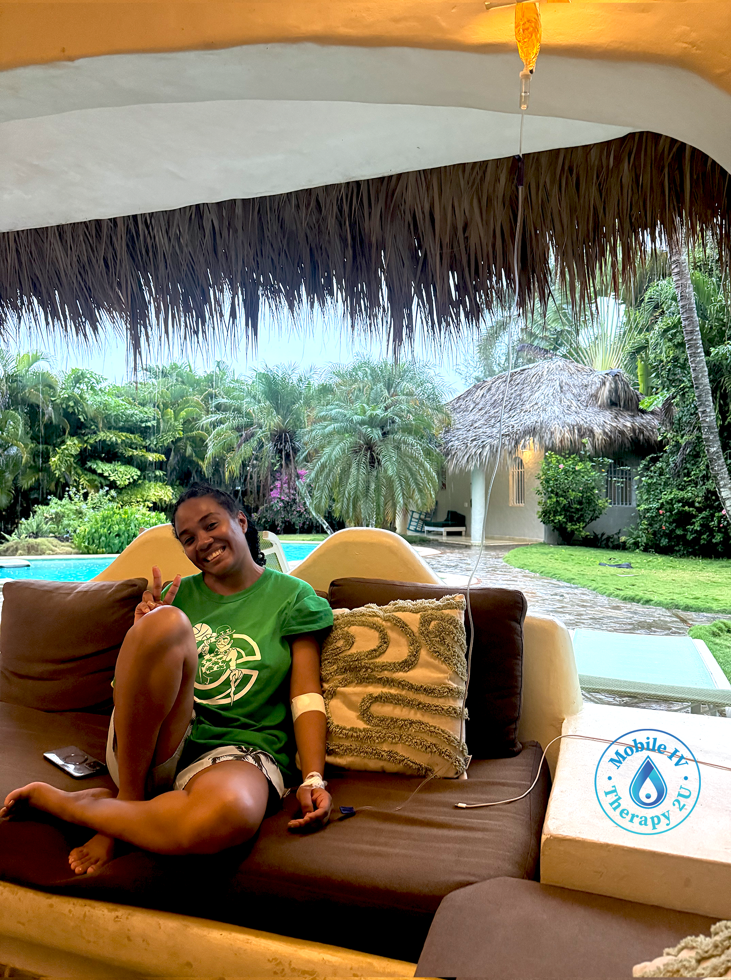 Young girl on holiday in an hotel, sitting on a sofa under a thatched roof, smiling and making a peace sign, with a tropical garden and swimming pool in the background, and a logo for Mobile IV Therapy in the bottom right corner.