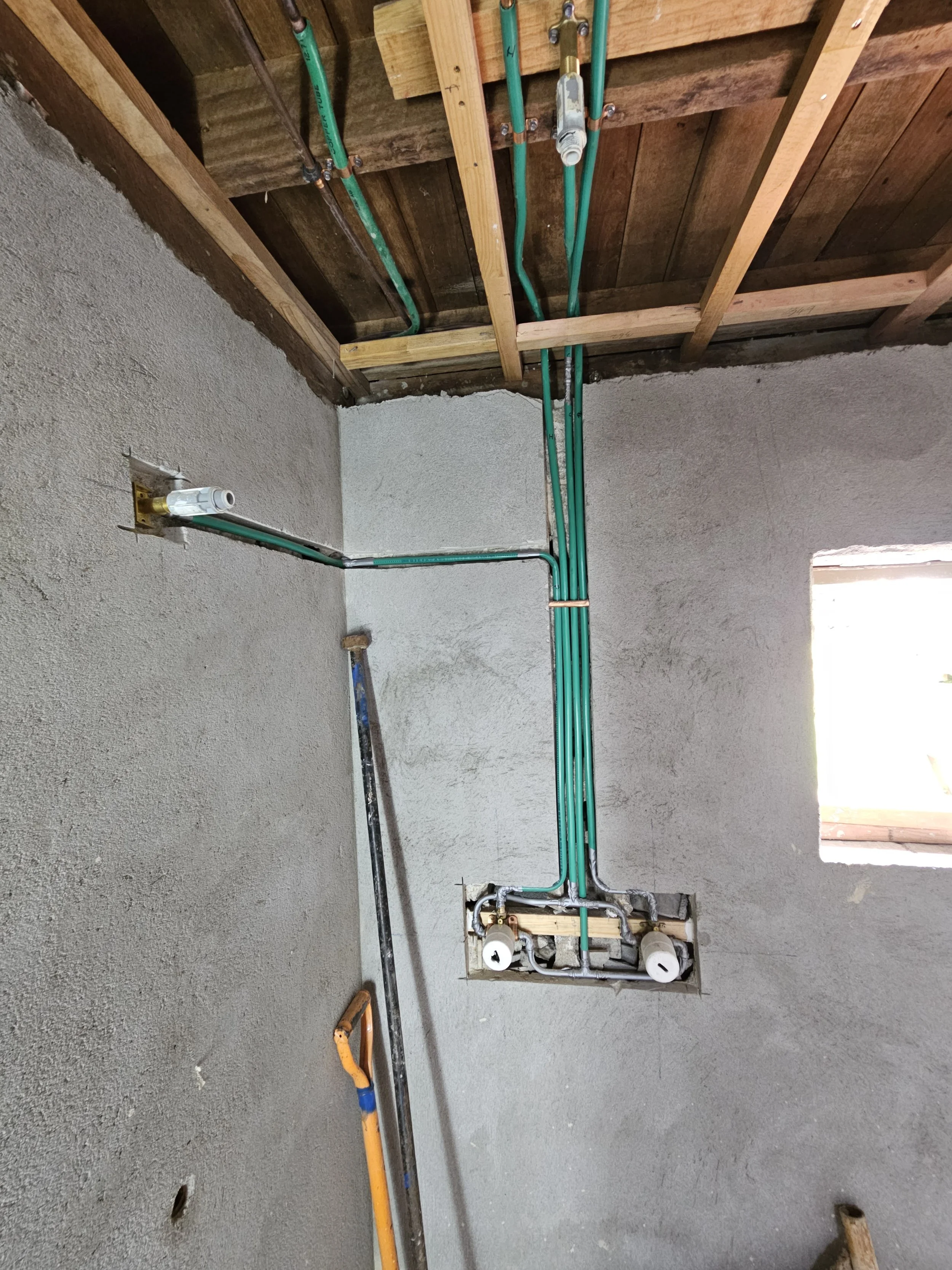 Installation of green lagged copper water pipes in a bathroom with a concrete wall and a wooden ceiling. Pipes enter through a wall opening and are held in place with fasteners. Tools leans against the wall.