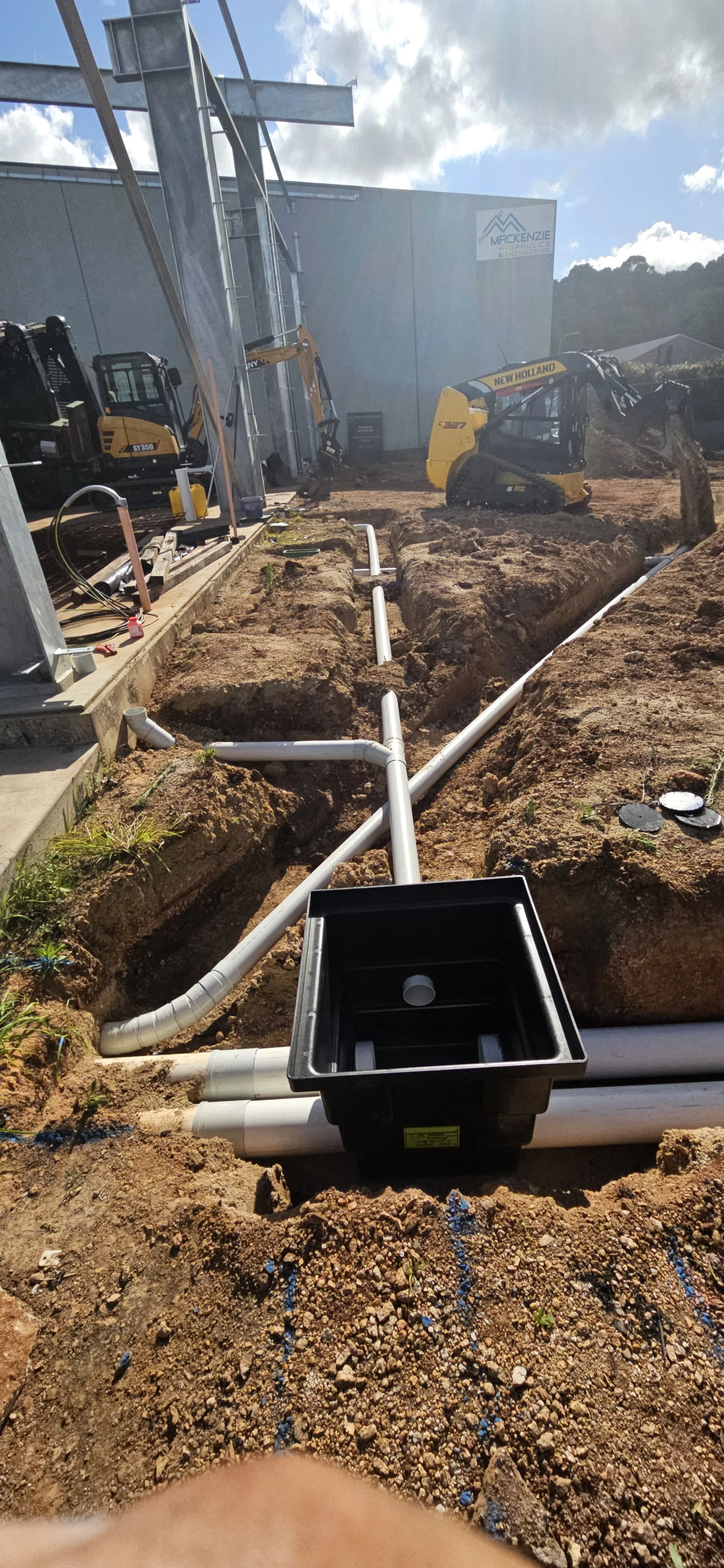 Construction site with exposed underground plumbing pipes and machinery, including mini excavators, next to a new industrial building.
