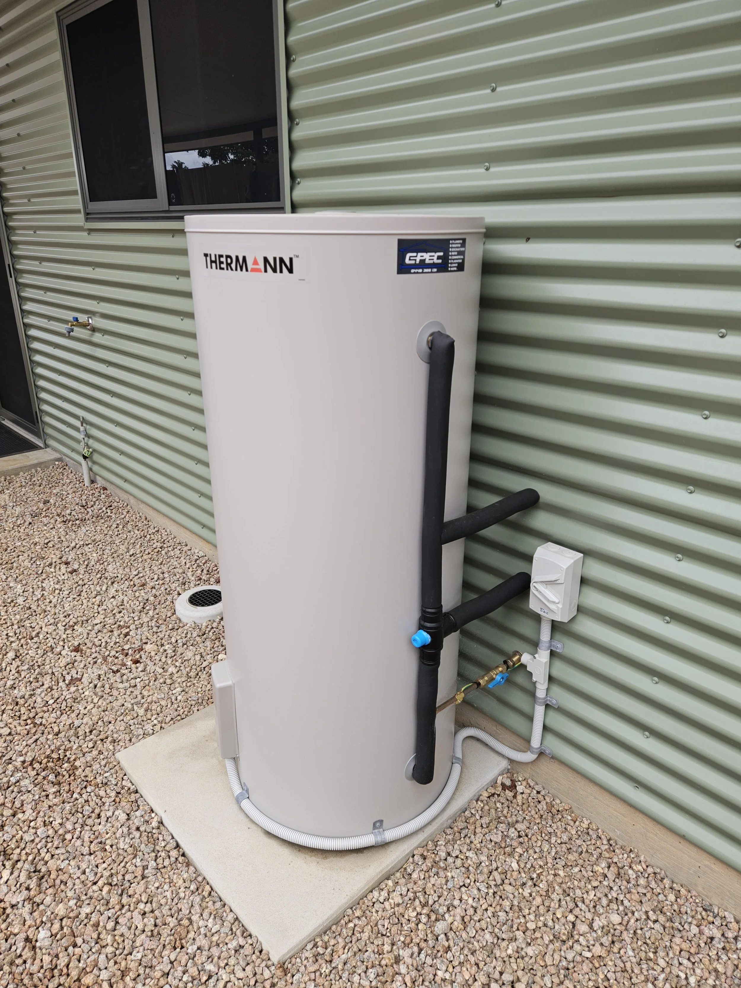 An outdoor water heater unit installed next to a green metal building wall, with black lagged water pipes and electrical connections visible, on a concrete pad surrounded by gravel.