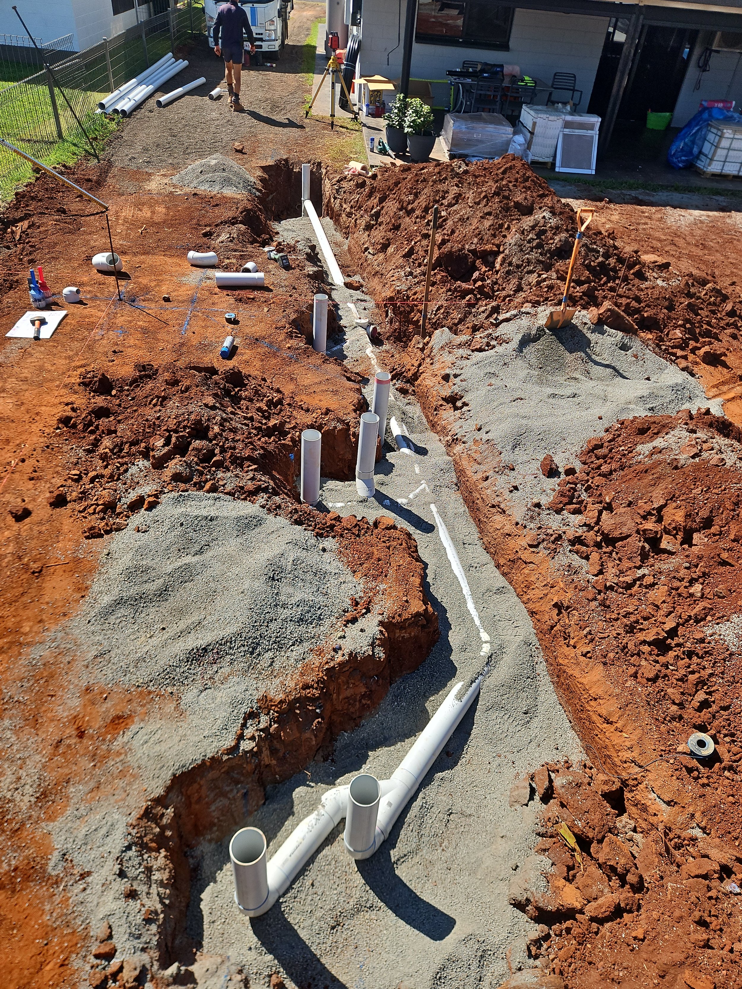 A construction site showing an area being prepared for underground piping. Several white PVC pipes are installed and labeled, with gravel and dirt around. Construction tools and materials are scattered nearby, and a worker and another individual are 