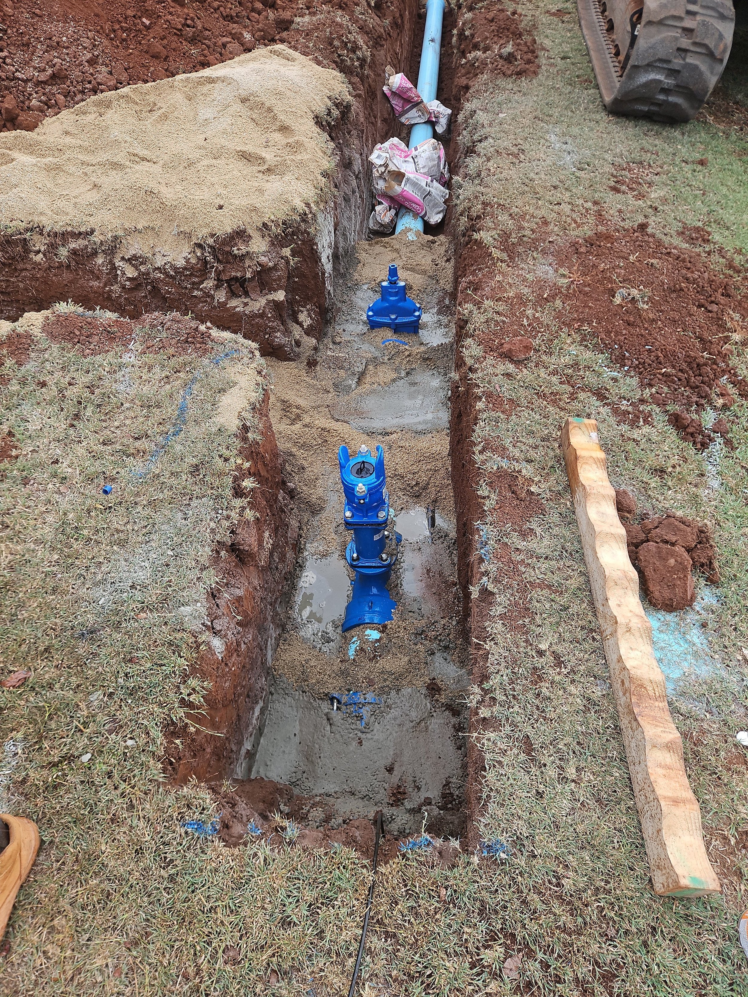 A trench with 150mm water mains valves and pipes being installed for water supply to a new subdivision. The trench is surrounded by grass and dirt, with construction tools nearby.