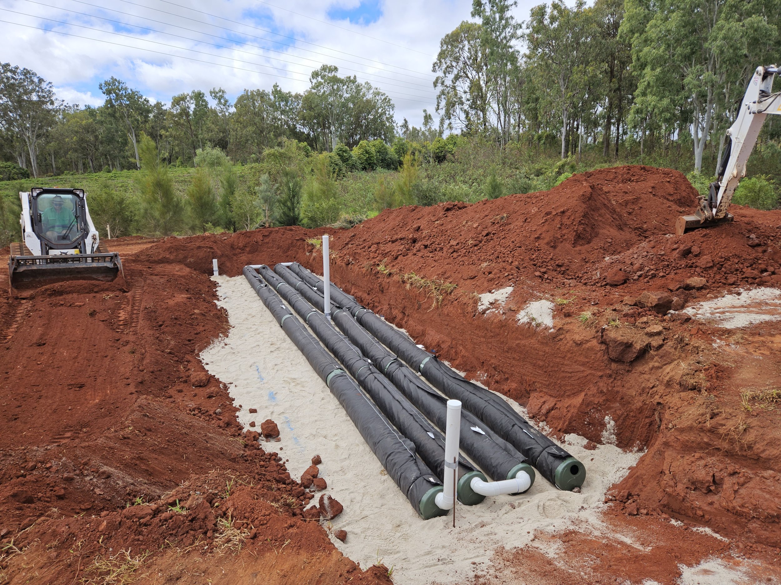 Construction site with AES septic pipes being installed in a trench, surrounded by red dirt and trees in the background.