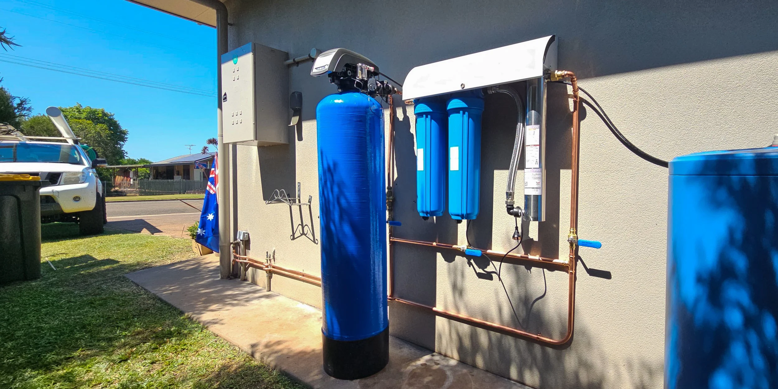 Outdoor water filtration system with blue tanks and copper pipes installed against a beige wall, next to a grassy area and parked vehicles.