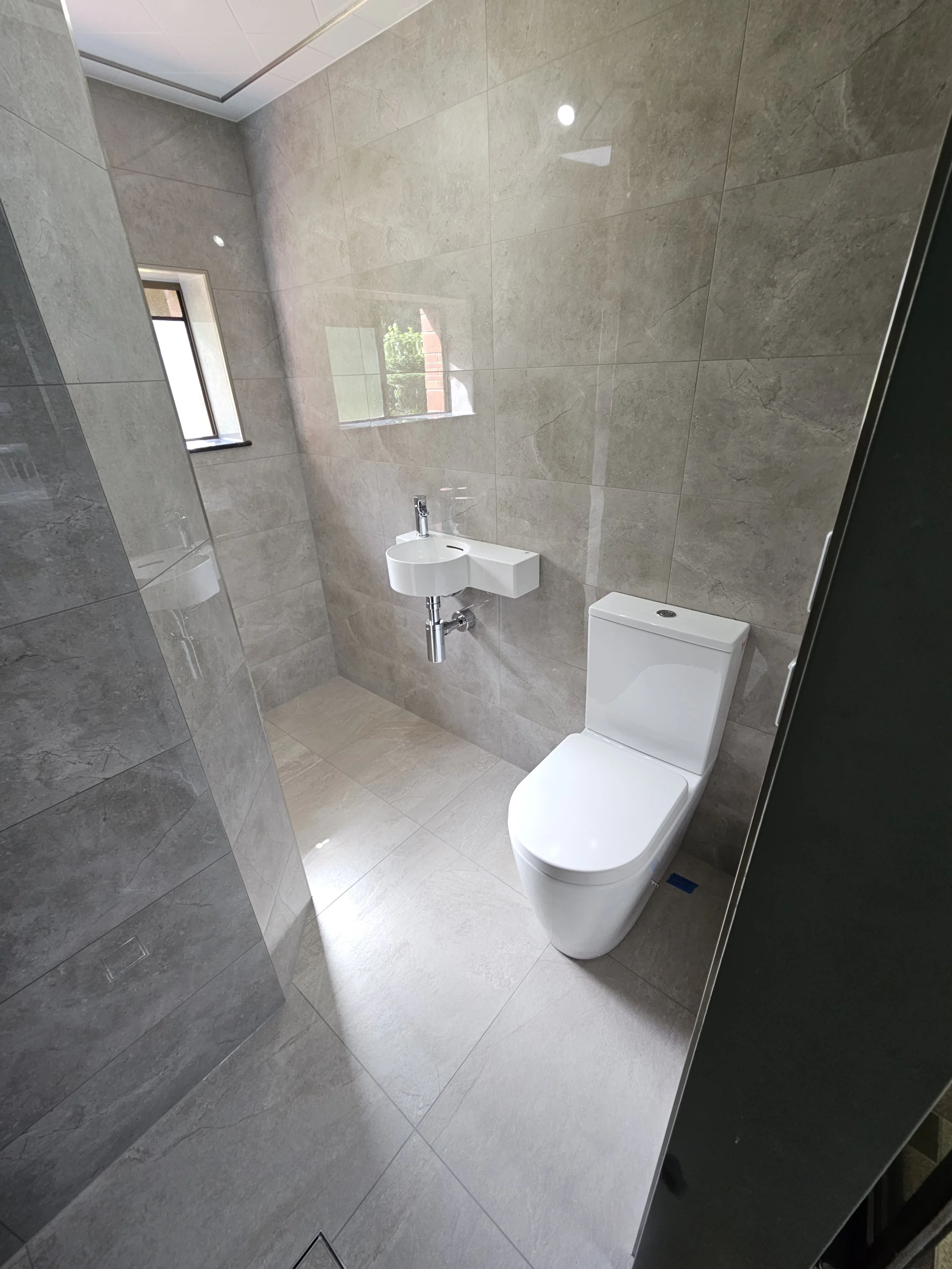 Modern bathroom with a white toilet, a small wall-mounted sink, and two small windows, featuring beige tile walls and floor.
