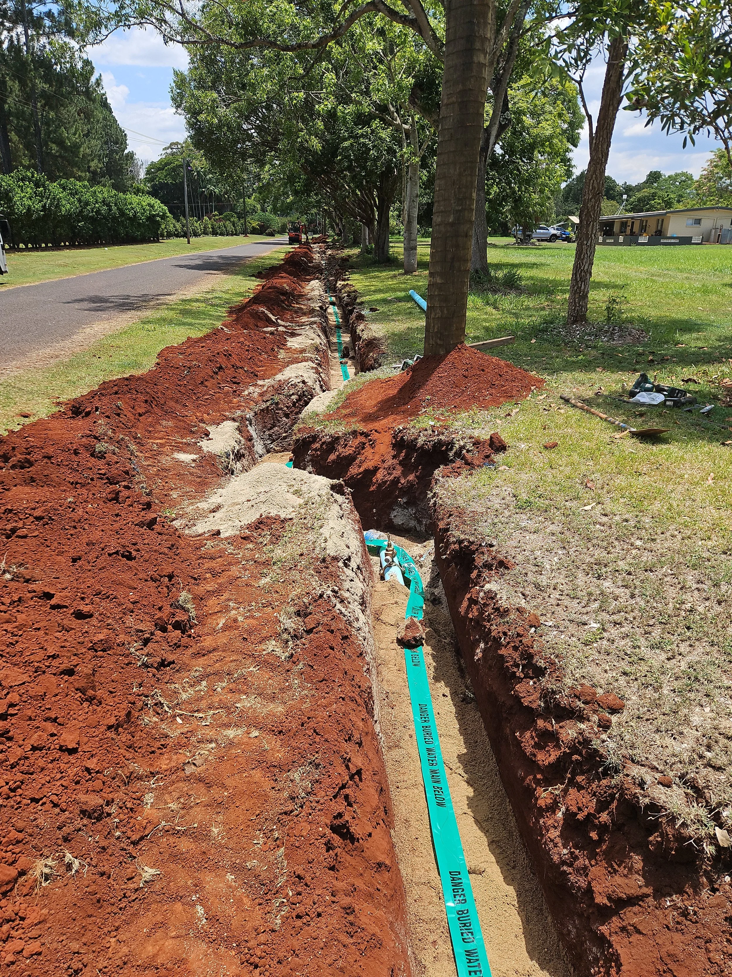 Construction workers installing or repairing underground utility pipes along a grassy roadside, with an open trench, trees, and vehicles in the background.