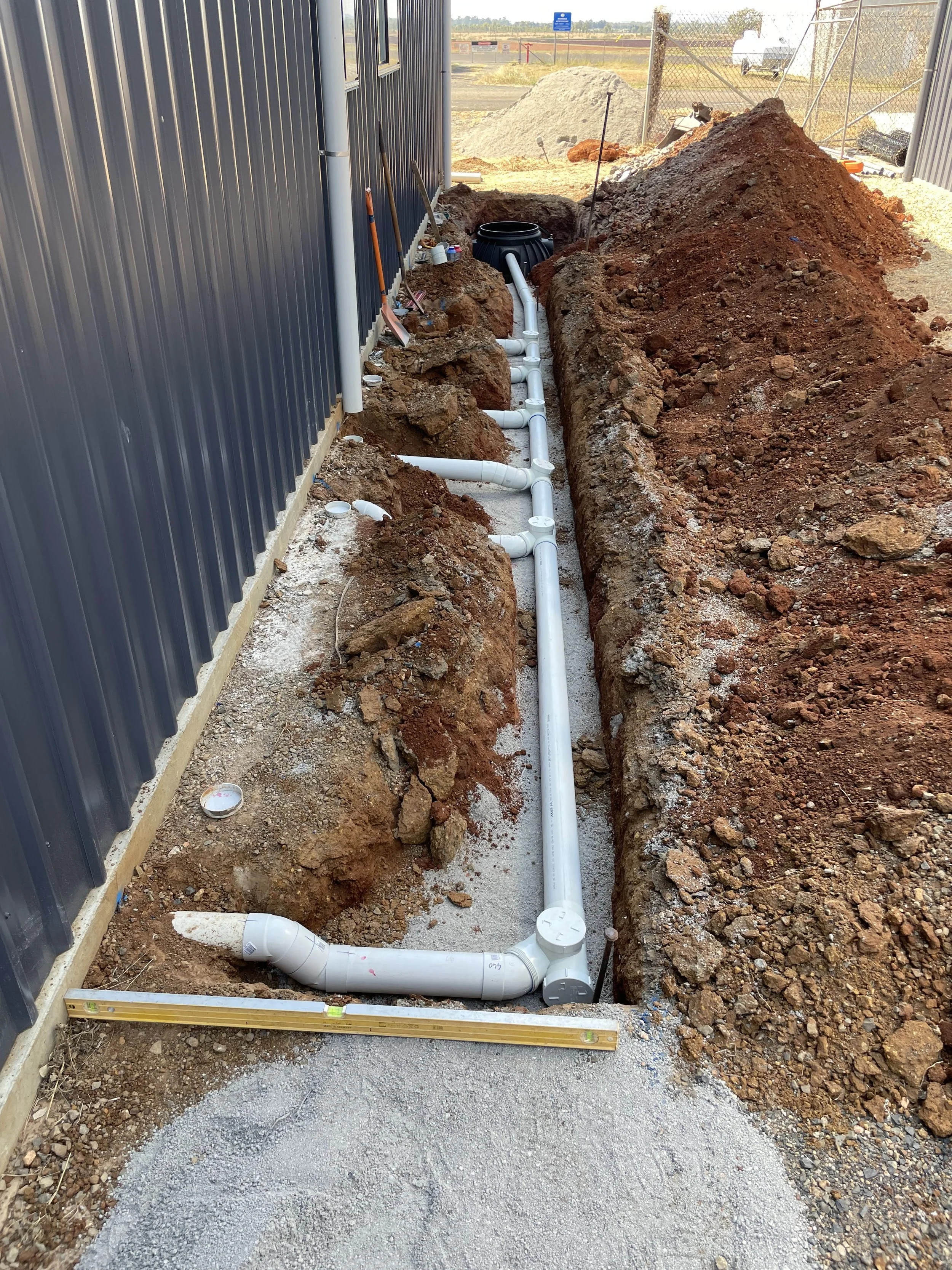 Construction site where new 100mm PVC sewer pipes are being installed underground along the side of a building, with a level tool resting on the ground nearby.