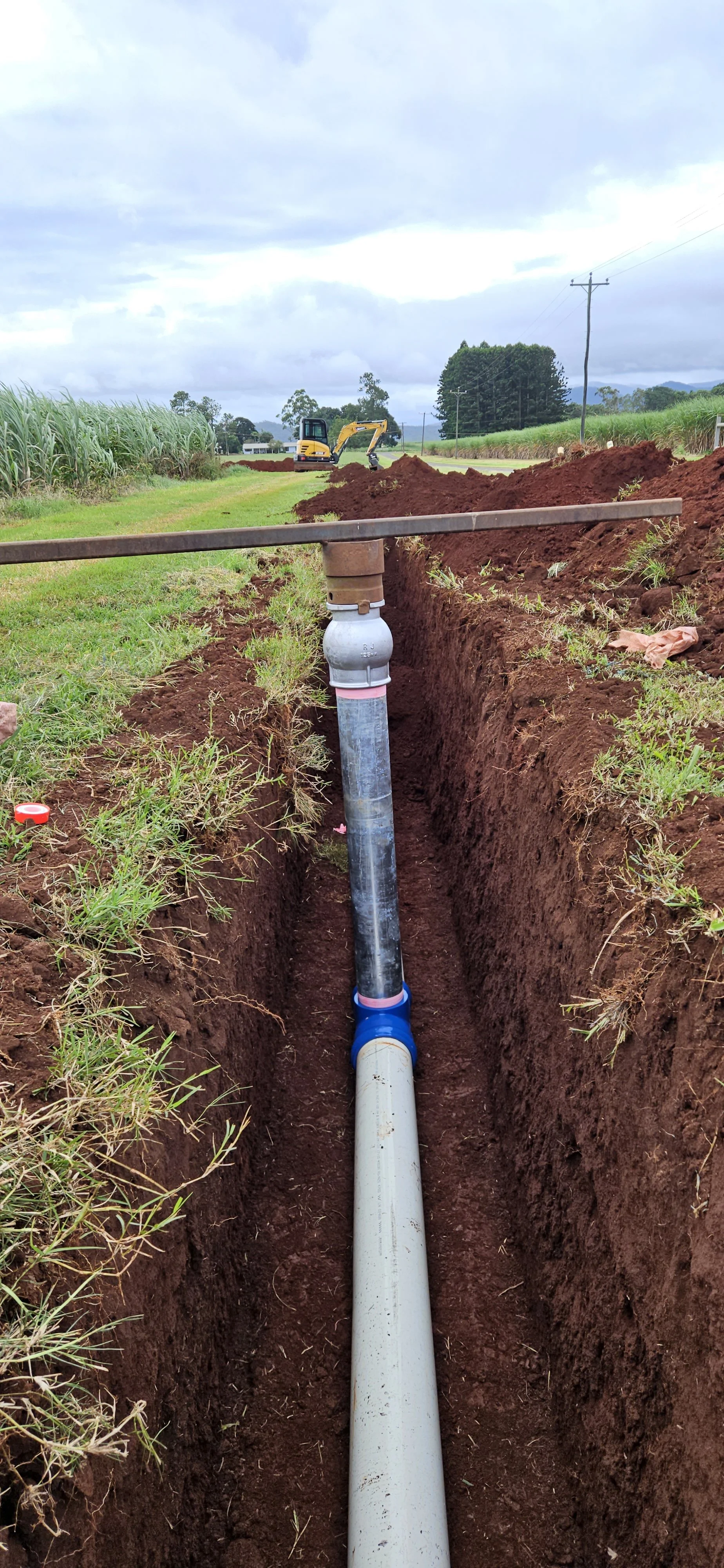 A trench with a large pipe being installed in rural area with grass, trees, overcast sky, and a small excavator in the background.