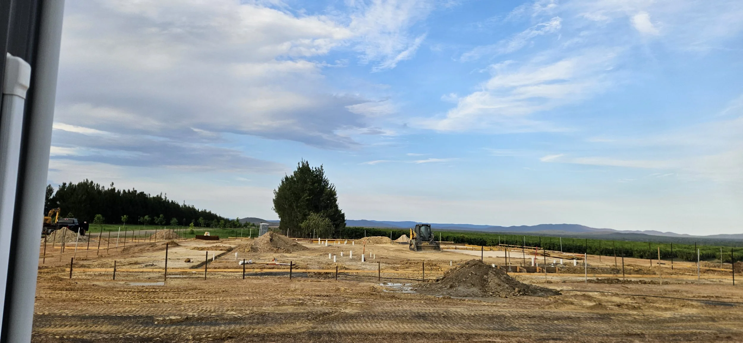 Construction site with dirt piles, small machinery, and pipes, with green fields, trees, and mountains in the background under a cloudy sky.