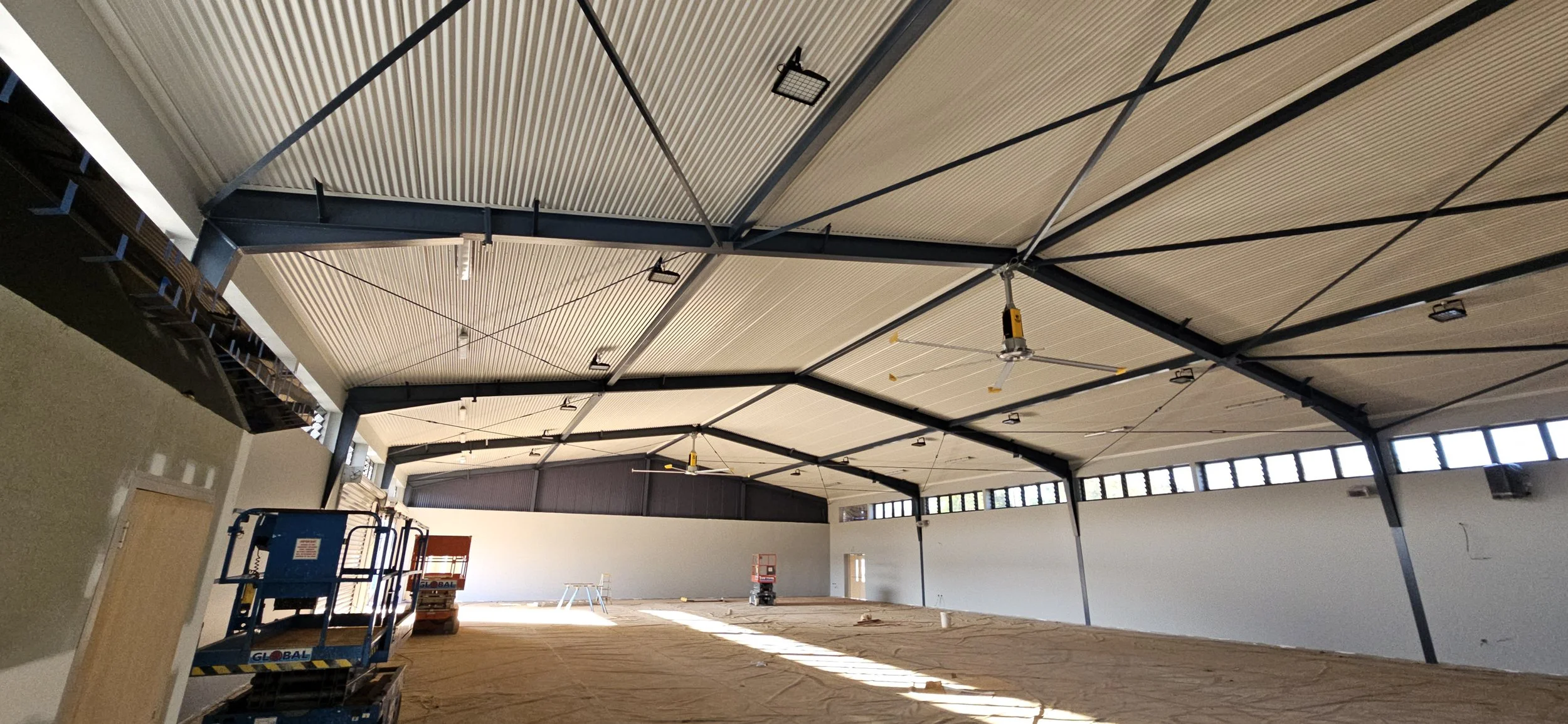 Interior of a large industrial building under construction with colorbond sheeting and flashings on a high ceiling, ceiling fans, and construction equipment.