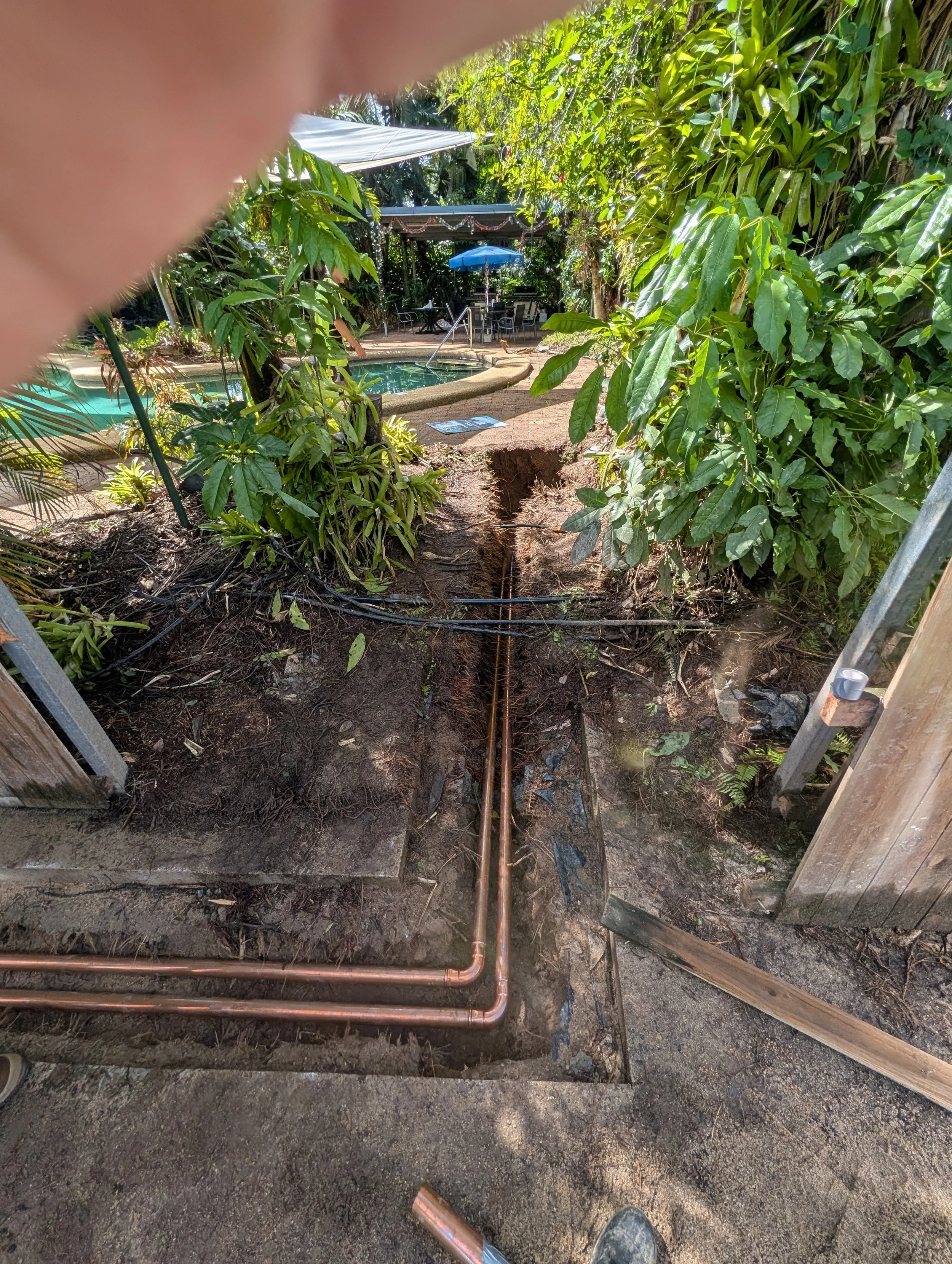 A unit complex near a pool with a freshly dug trench and new copper pipes installed in the ground, surrounded by lush green plants and outdoor furniture with umbrellas.