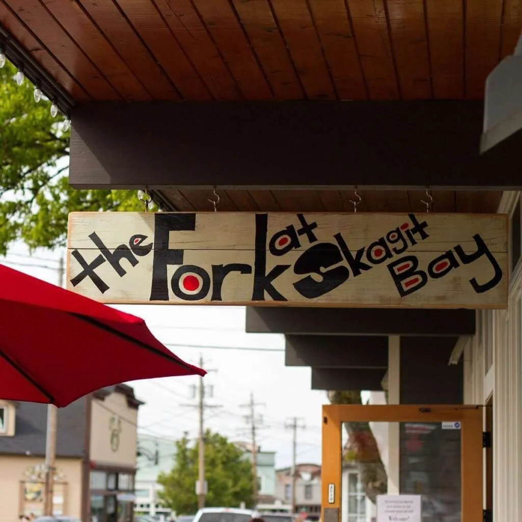 Wooden sign hanging outside a building reading 'the Fork at Skagit Bay' with a red umbrella partially visible on the left and trees in the background.