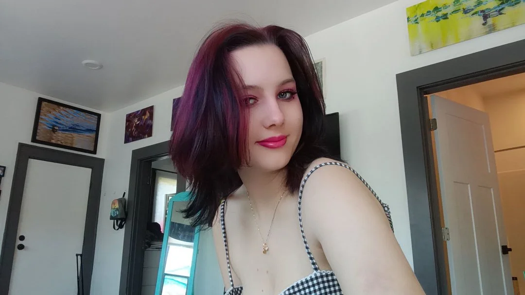 Young woman with shoulder-length dark hair and reddish-purple highlights, wearing a checkered spaghetti strap top, smiling in a well-lit room with artwork on the walls.