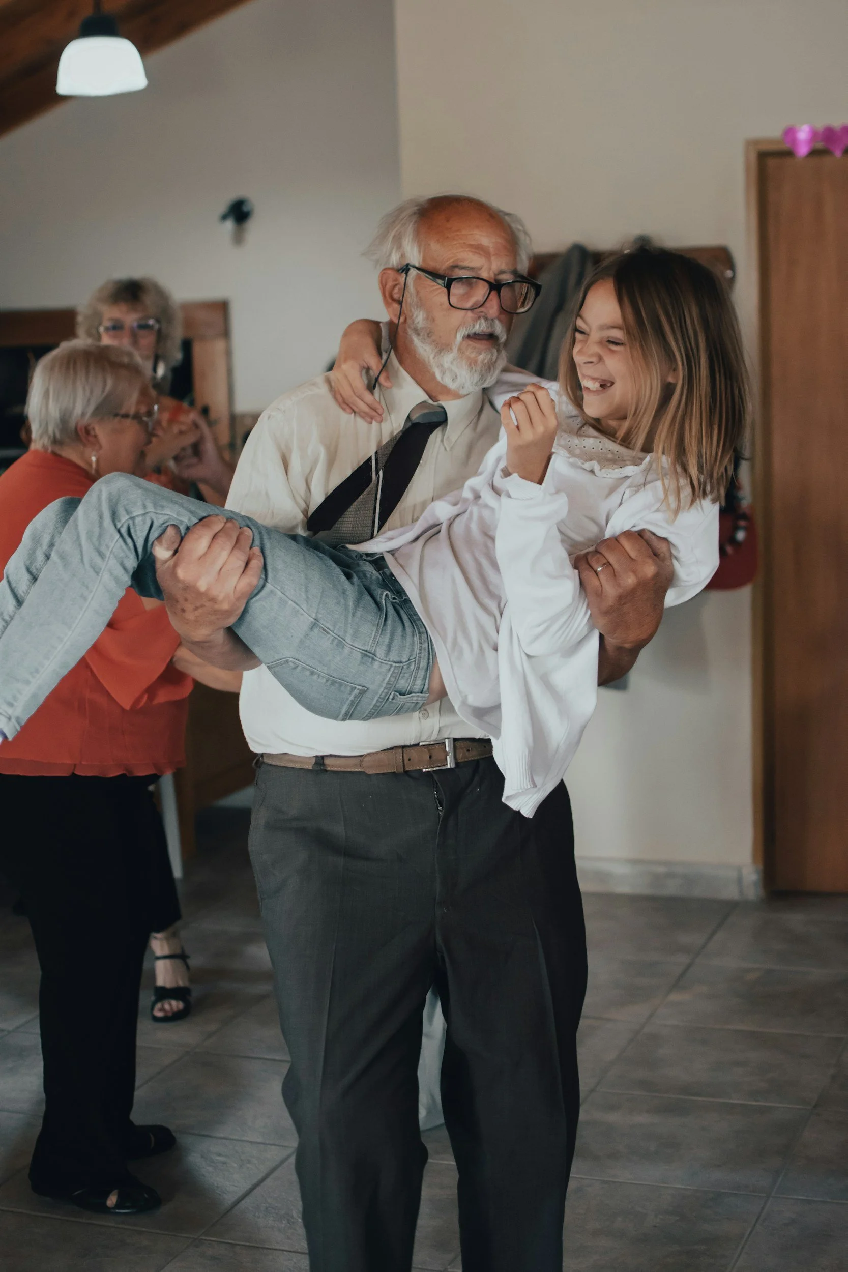 An elderly man with glasses and a white beard is dancing while holding a young girl with long hair. The girl is laughing and wearing a white dress. In the background, two women are also seen, one wearing an orange top and glasses. The scene takes place indoors, with a wooden ceiling and tiled floor.
