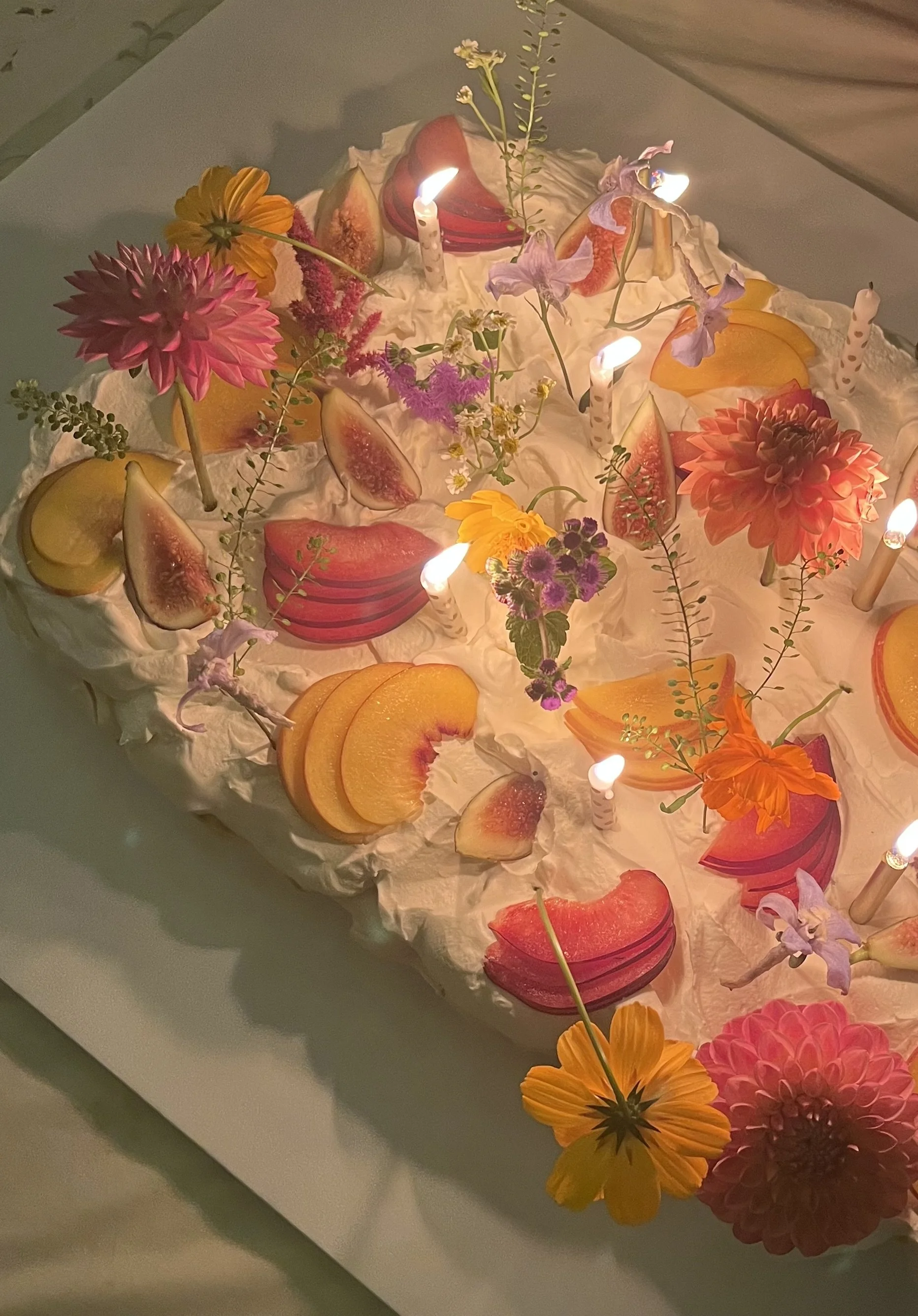 A colorful cake decorated with sliced peaches, figs, and fresh flowers, with lit candles on top.