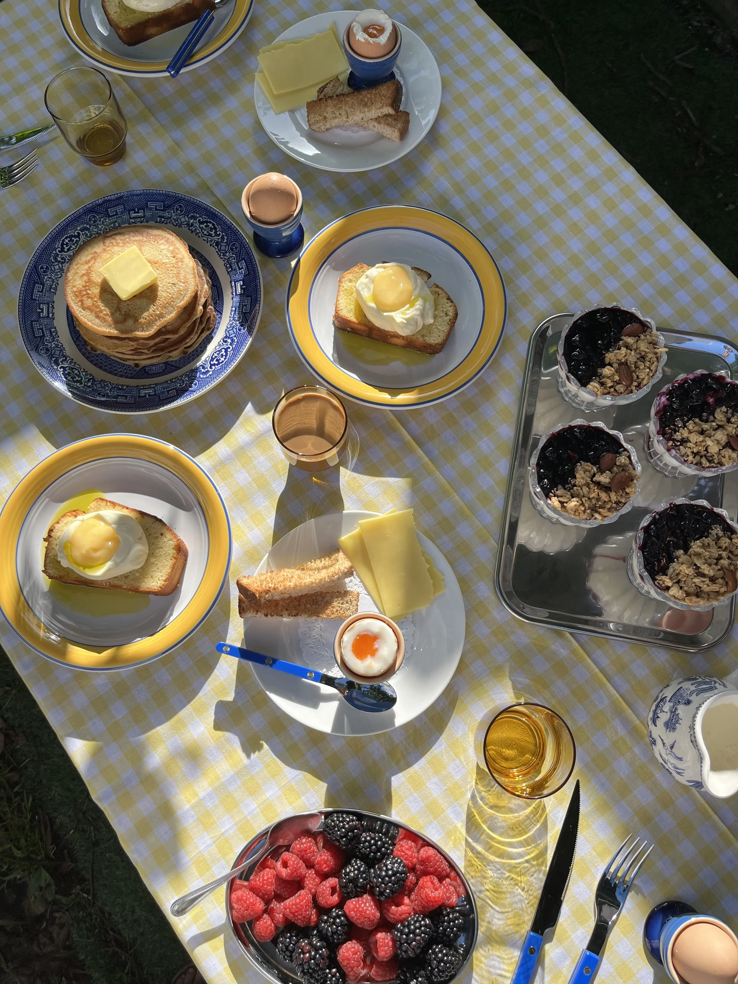 A breakfast table with pancakes topped with butter, eggs on toast, slices of cheese, bread, a bowl of mixed berries, granola yogurt cups, and glasses of juice, set on a yellow and white checkered tablecloth.
