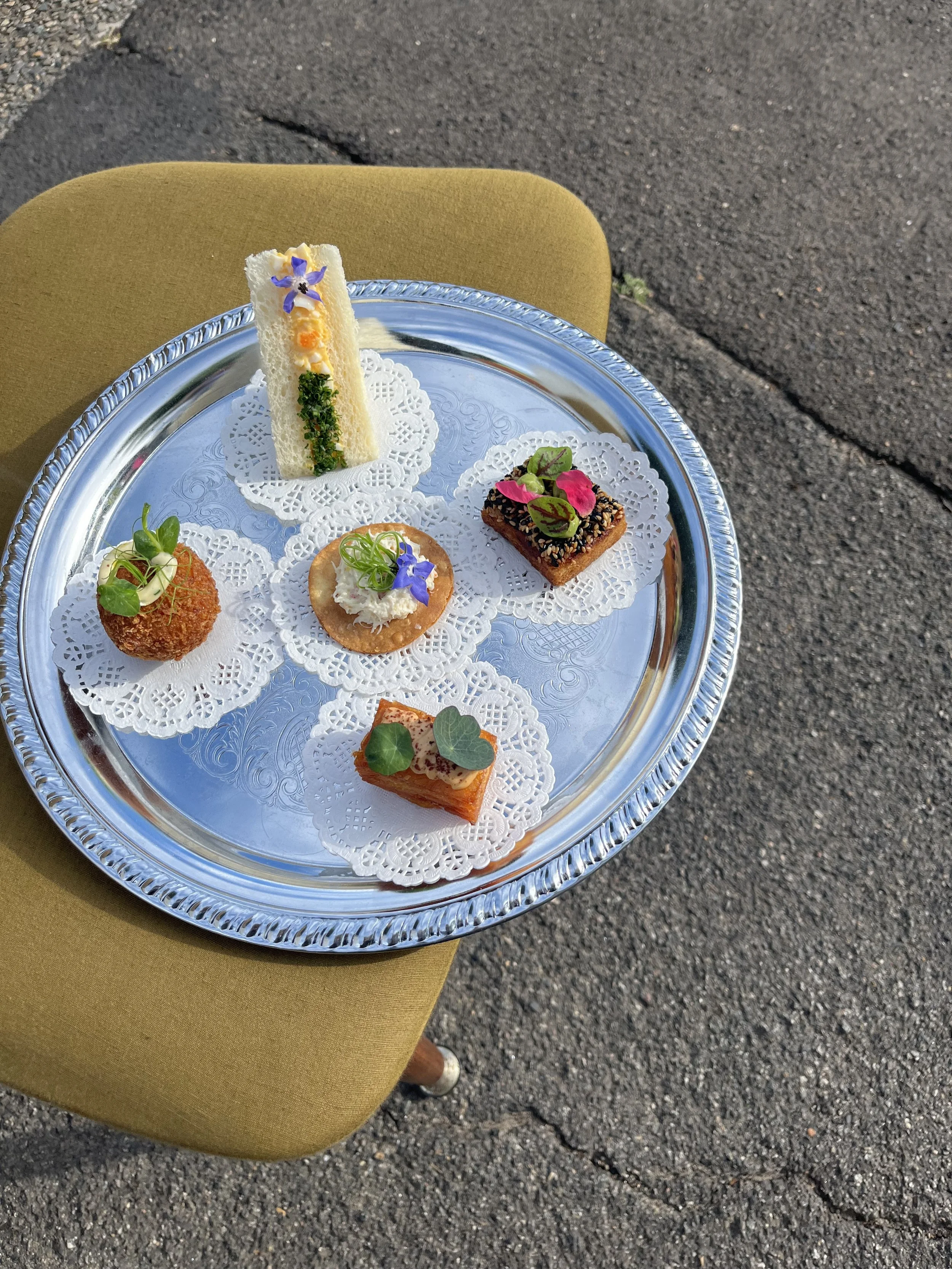 A silver tray with several colorful appetizers, including sandwiches, and garnished with edible flowers and greens, resting on a yellow cushion outdoors.